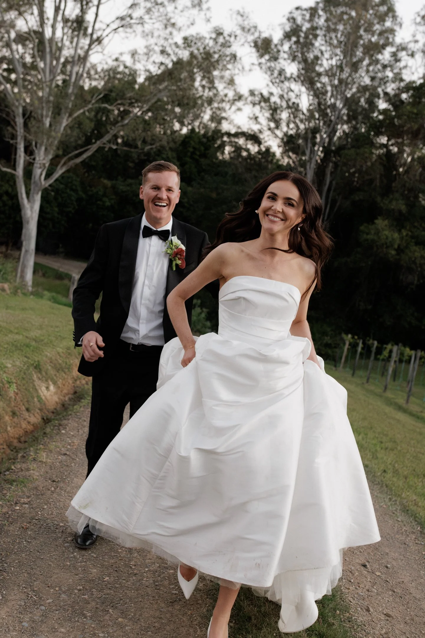 A smiling bride in a white wedding gown and a groom in a tuxedo with a bow tie, running outdoors on a dirt path, with trees in the background.