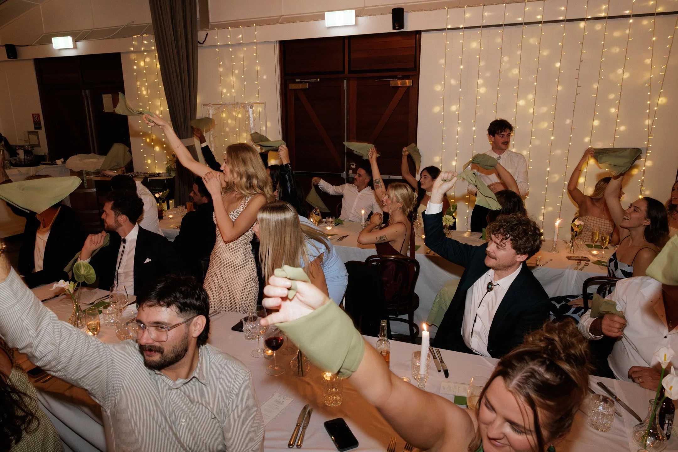 People at a celebration event sitting at tables, waving napkins, with fairy lights hanging on the wall in the background.