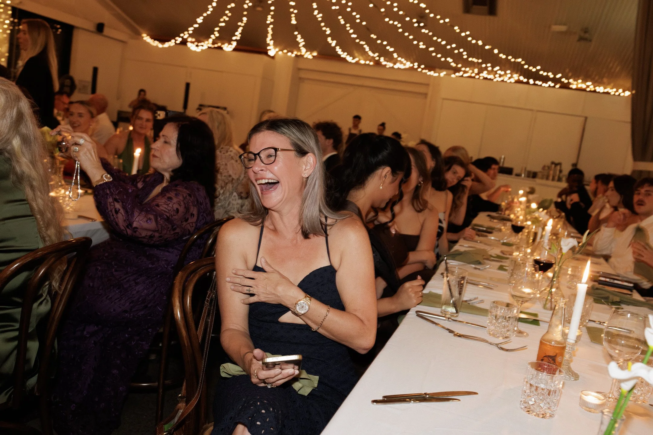 A group of people enjoying a celebration at a long dinner table decorated with candles and floral arrangements, with string lights hanging from the ceiling.