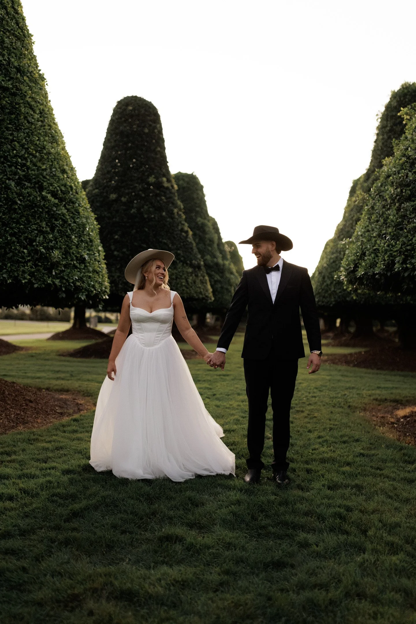 A bride and groom holding hands and smiling at each other outdoors, surrounded by tall, neatly trimmed trees and a green lawn.