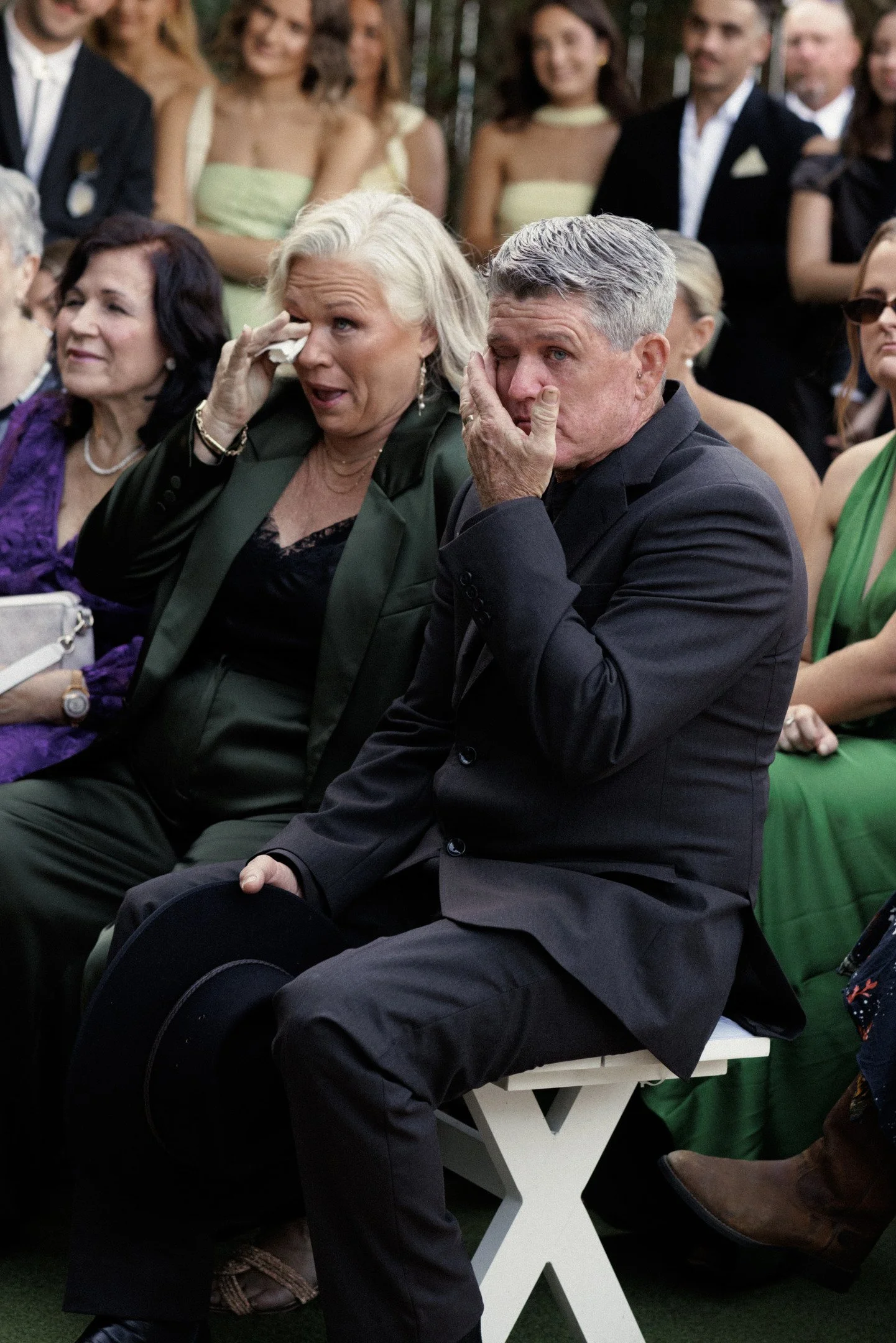 A group of people, mostly women, appear emotional at a formal event. The two older individuals in the foreground are wiping tears from their eyes, with some holding tissues. The background includes younger adults dressed in formal attire, smiling and