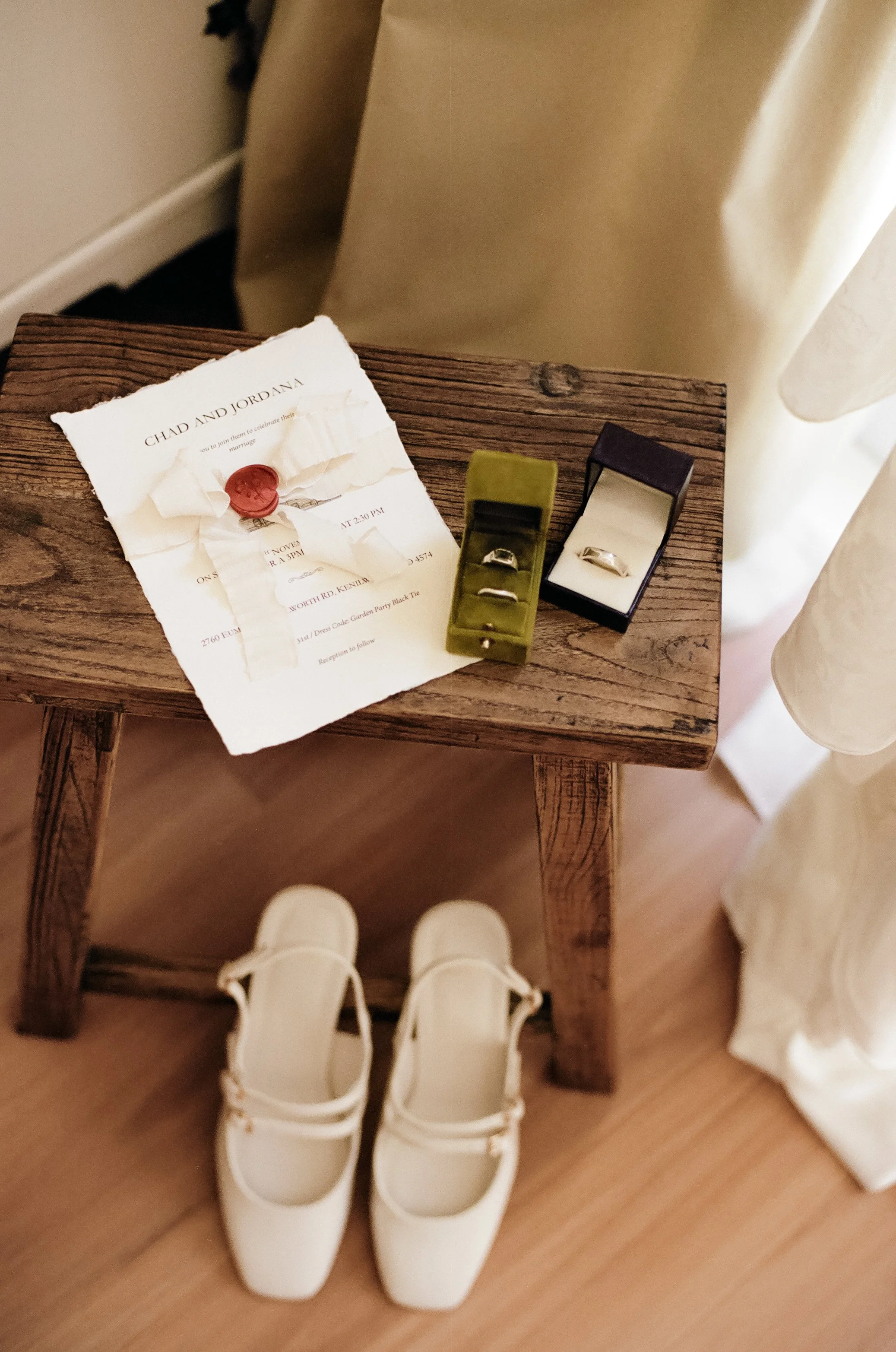 Wedding rings on a box and a paper receipt, white shoes below on a wooden floor, and a small wooden table with a wedding invitation and boxes for rings.