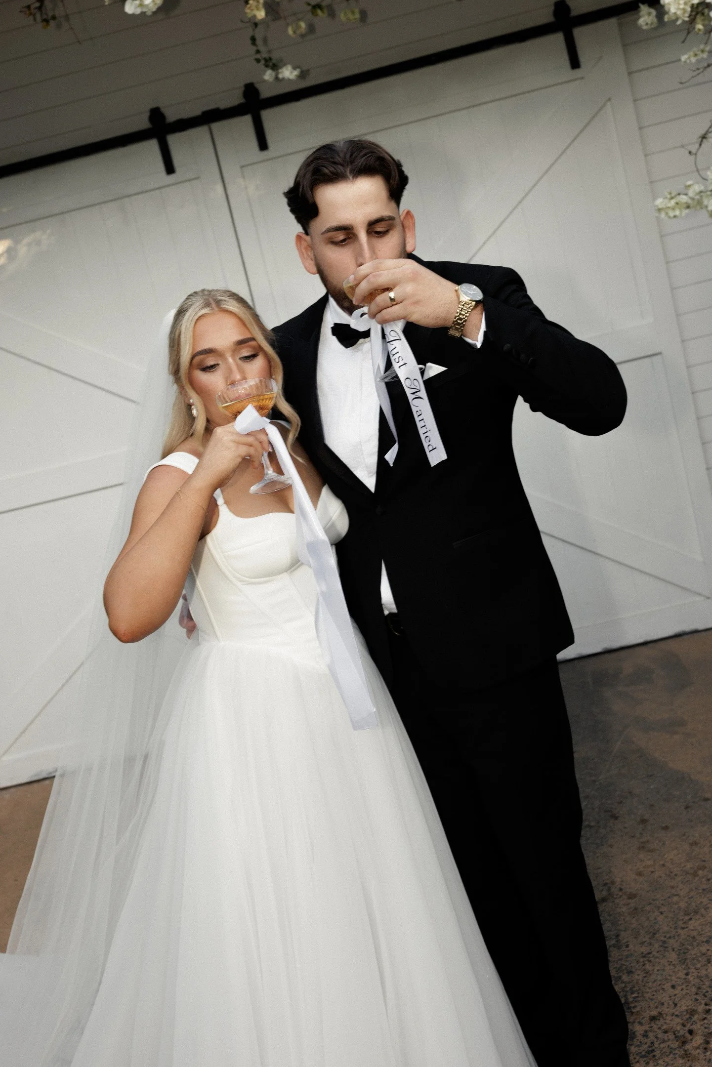 A bride and groom at their wedding, wearing formal attire and drinking from champagne glasses, standing inside a decorated venue.