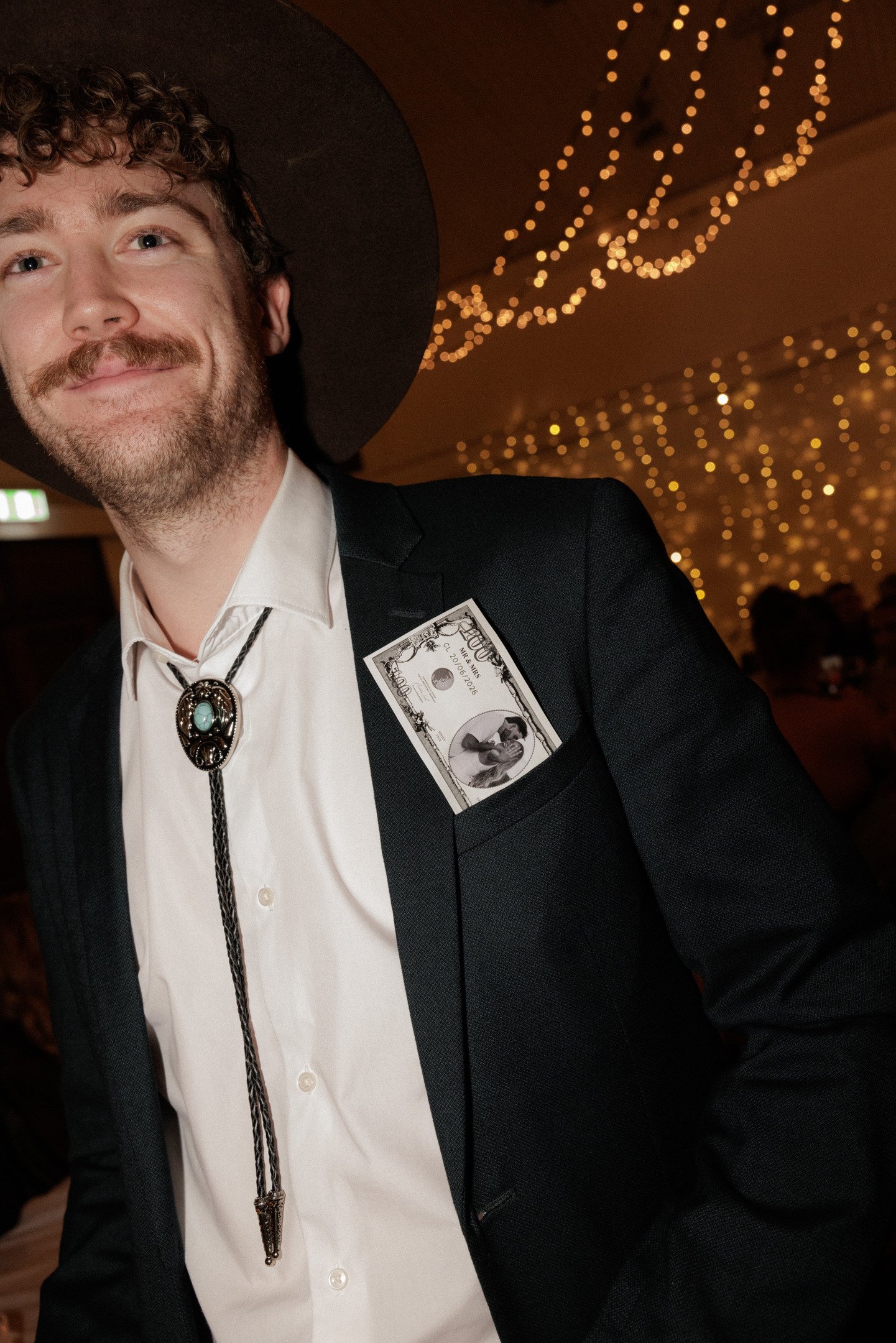 A man dressed in Western attire with a bolo tie, who is wearing a black blazer with a dollar bill tucked into the pocket, smiling at a social event with warm, fairy lights in the background.
