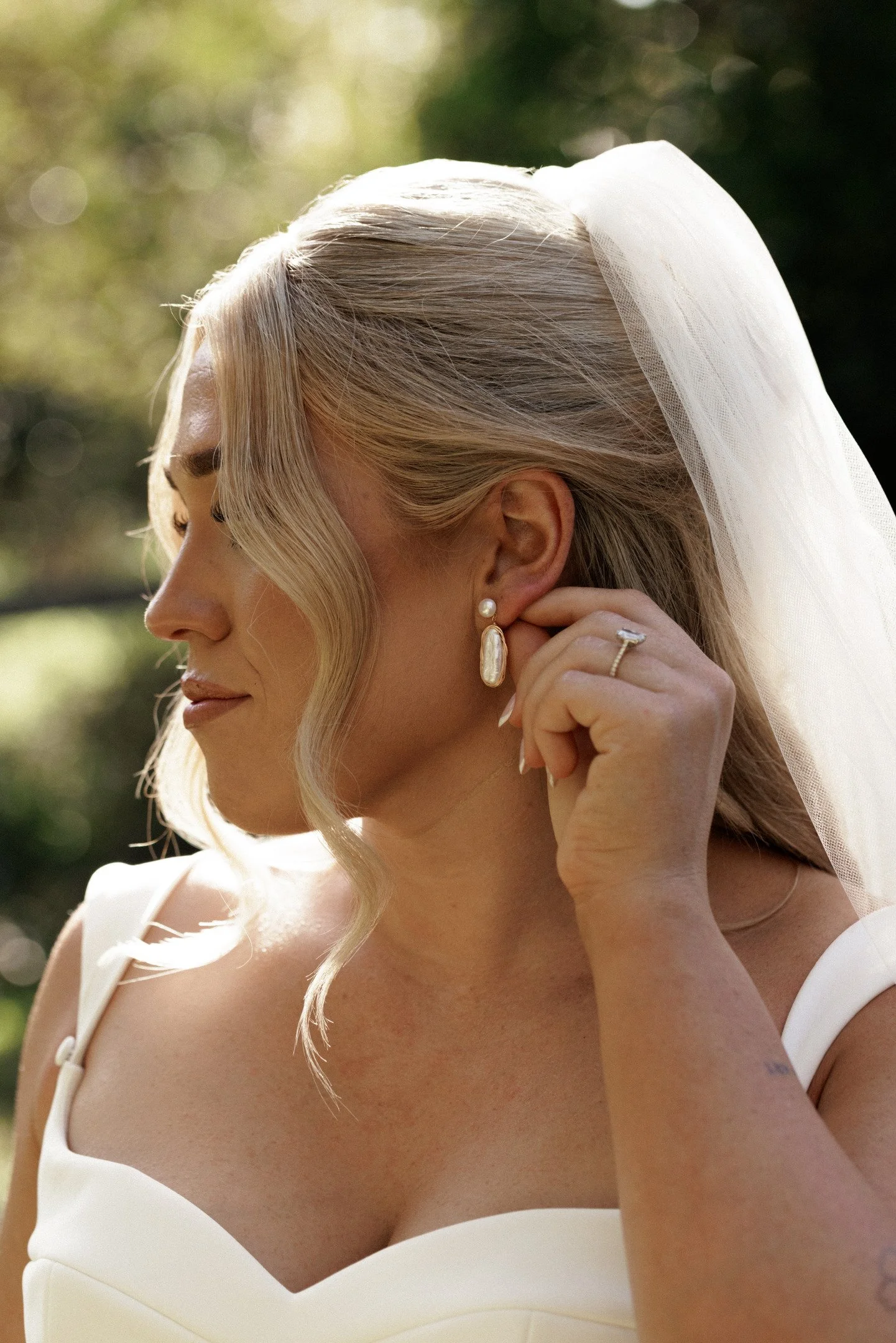 A woman with blonde hair styled in loose curls wearing a wedding dress and veil, with pearl earrings. She is touching her ear and rings, with soft sunlight and blurred greenery in the background.