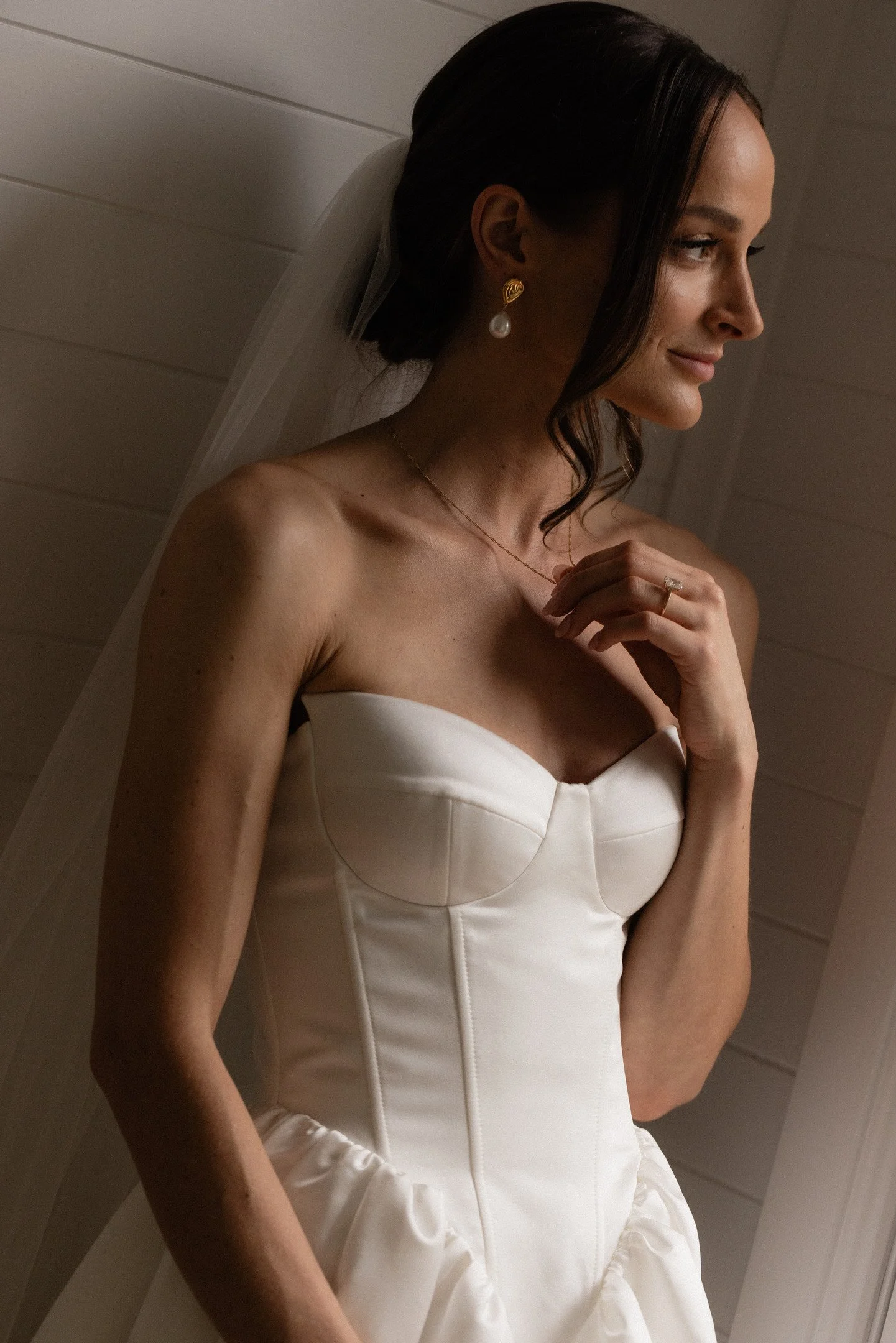 A woman in a white strapless wedding dress with dark hair styled in loose waves, wearing pearl earrings, a thin necklace, and a wedding ring, standing against a wood-paneled wall.