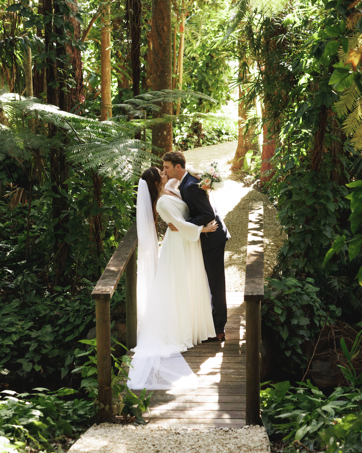 A bride and groom sharing a kiss on a small wooden bridge surrounded by lush green tropical plants and trees.