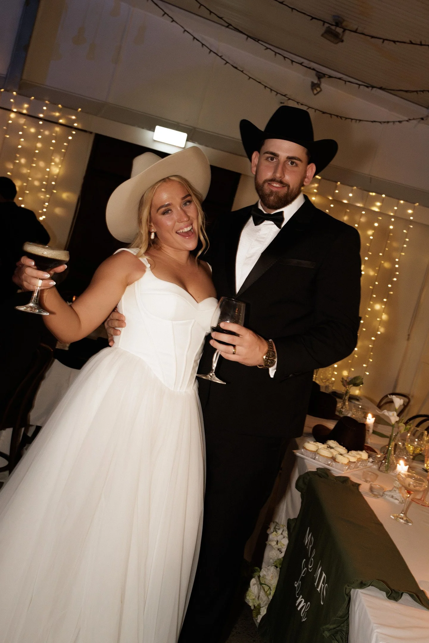 Man and woman dressed in wedding attire, wearing cowboy hats, holding drinks, smiling at a celebration or reception with string lights in the background.