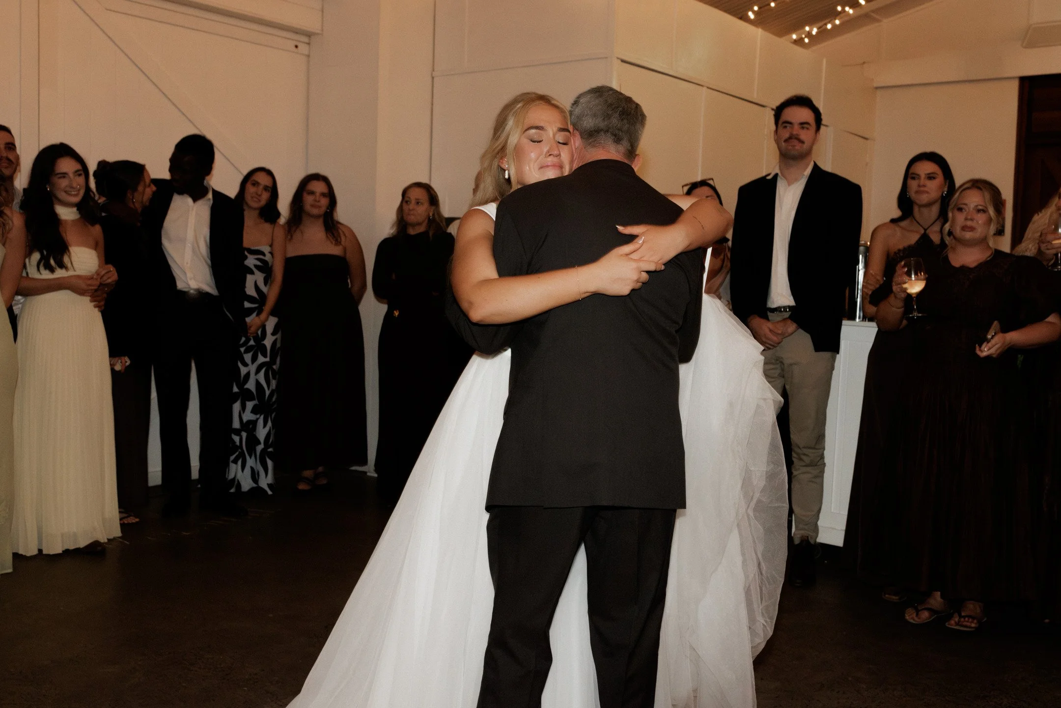 A bride and groom share a dance at their wedding reception, surrounded by guests watching and smiling.