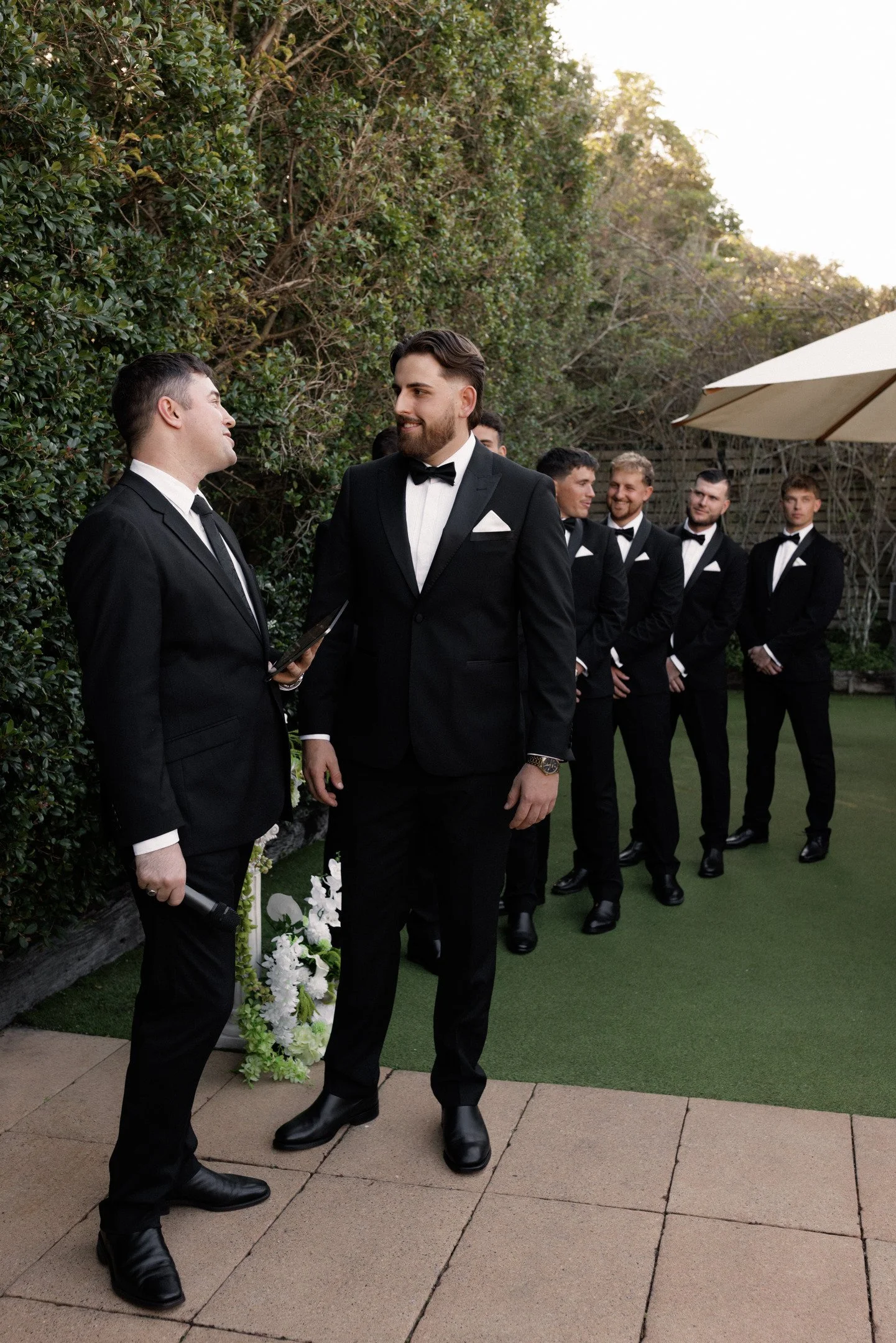 Groom and groomsmen dressed in black tuxedos with bow ties, standing outdoors on a paved patio with ivy-covered wall and hedge in the background, during a wedding celebration.