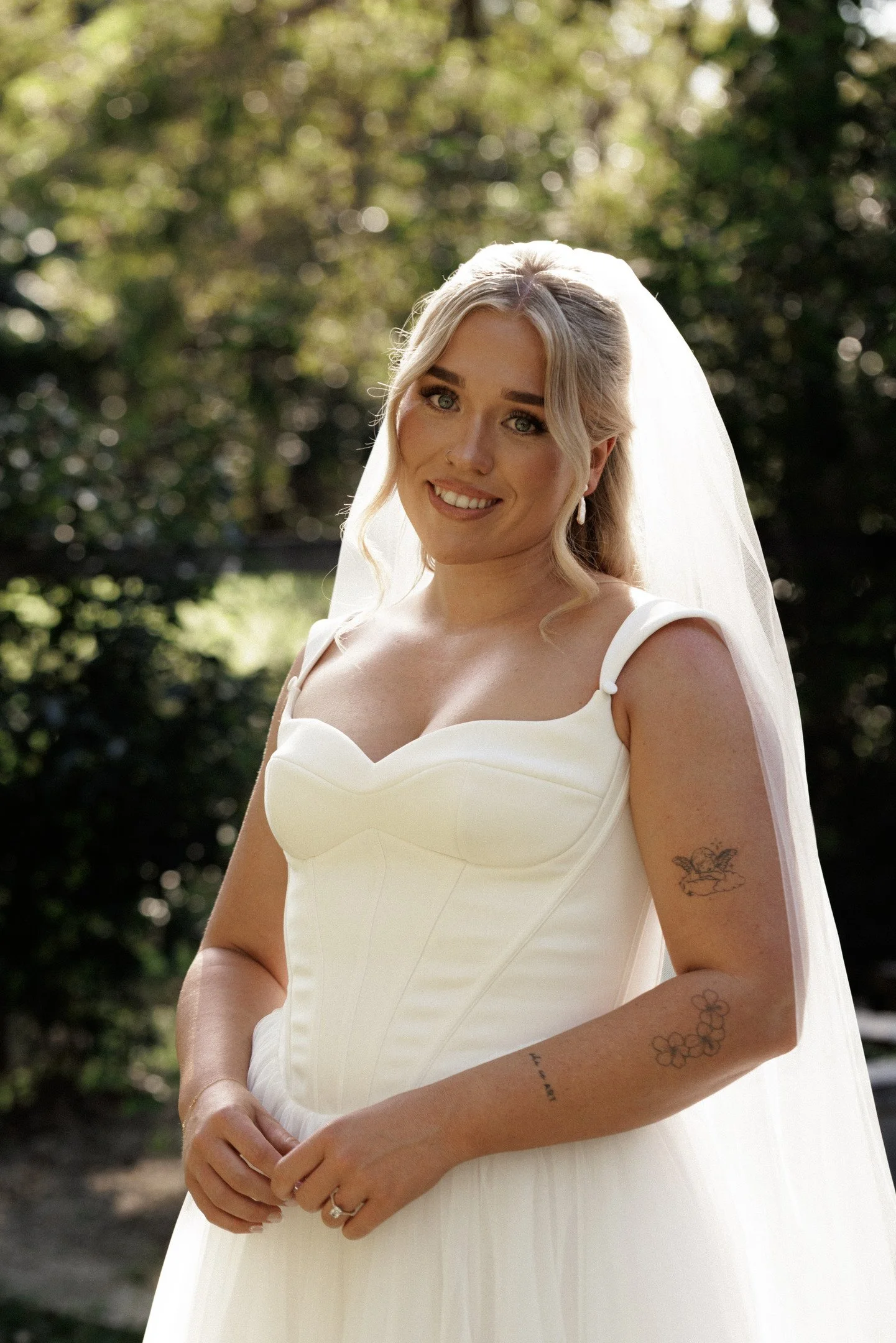 A bride with blonde hair and blue eyes, wearing a white wedding dress with shoulder straps and a veil, standing outdoors with blurred greenery in the background.