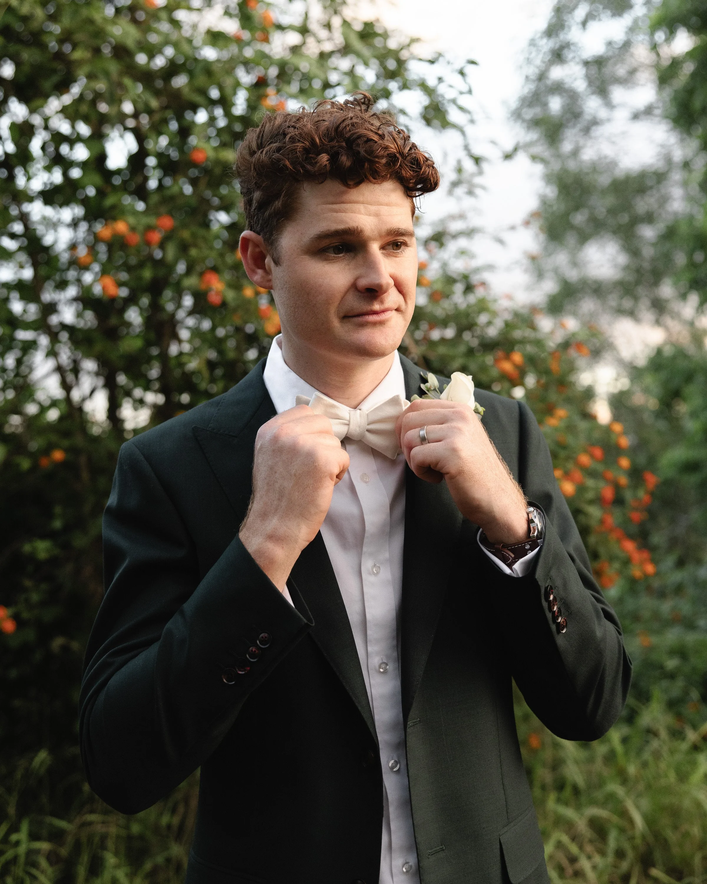 Man adjusting bow tie, wearing a suit in a garden setting with green foliage and orange flowers -Elevated Weddings