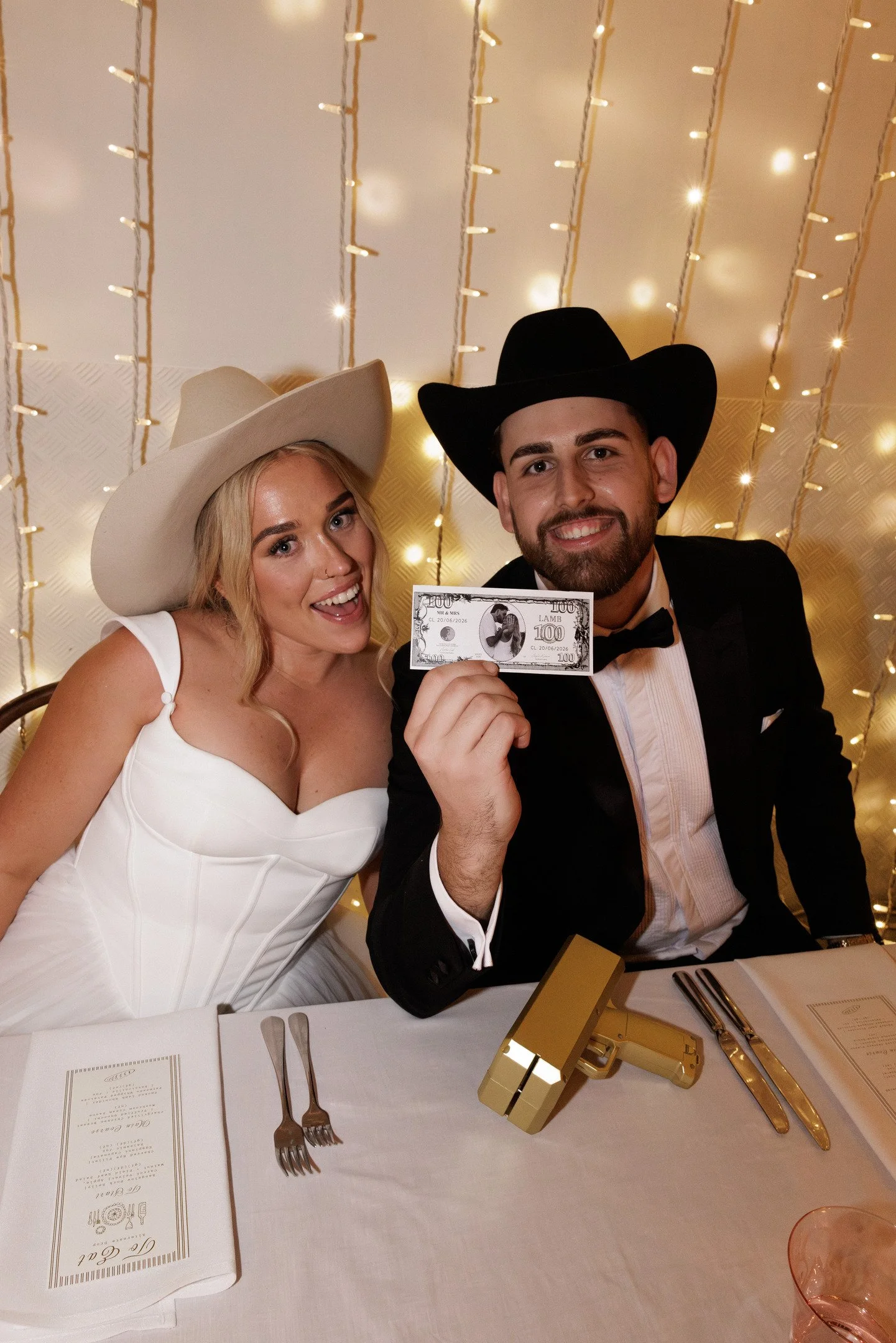 A smiling couple dressed in formal attire at a celebration, with the woman in a white dress and wearing a large cowboy hat, and the man in a tuxedo and wearing a black cowboy hat. The background has string lights, and they are holding a prop resembli
