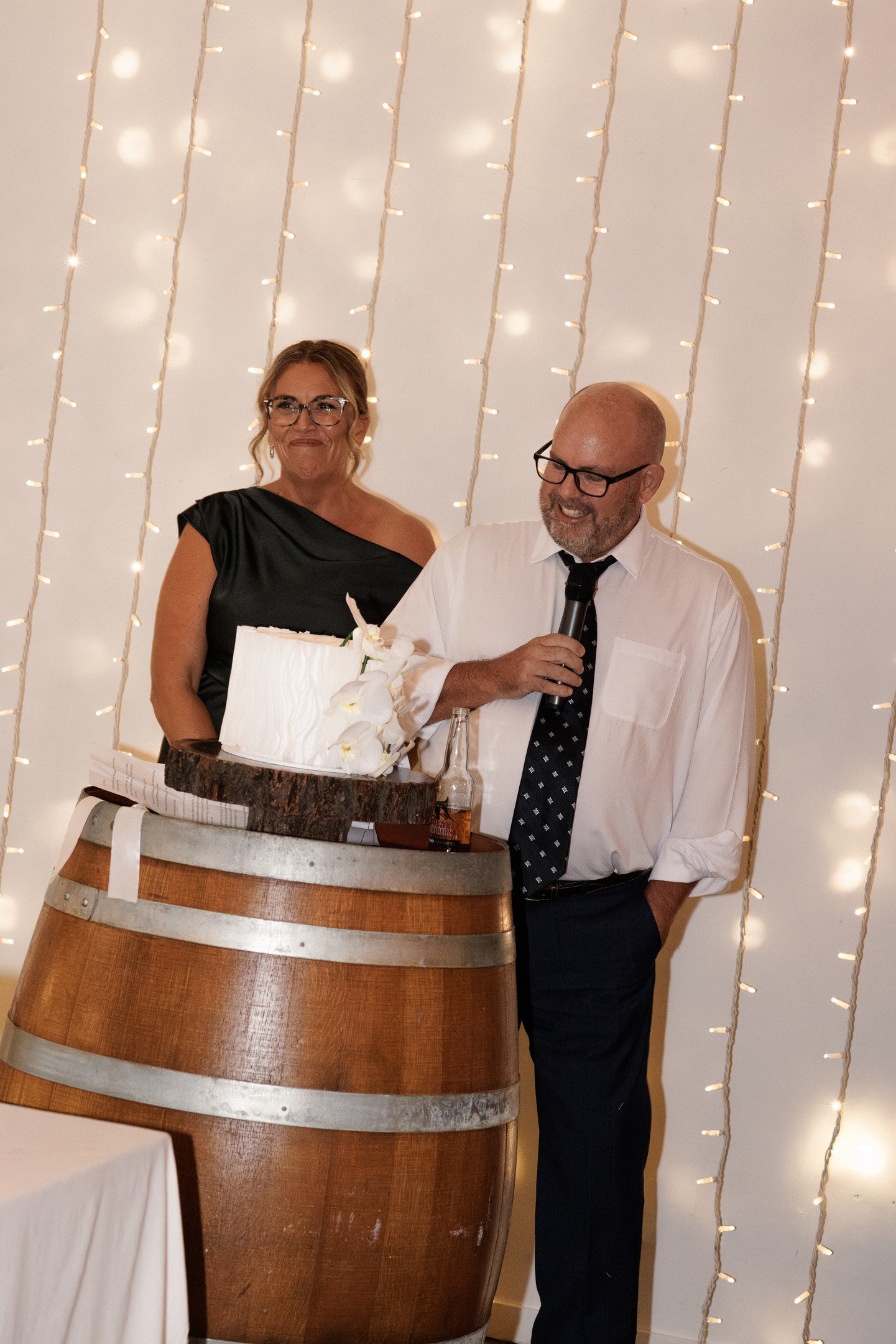 A man in a white dress shirt, black tie, and glasses is holding a microphone and smiling as he stands near a woman in a black dress, who is also smiling. They are behind a large wooden barrel with a white cake on top, decorated with flowers. The back