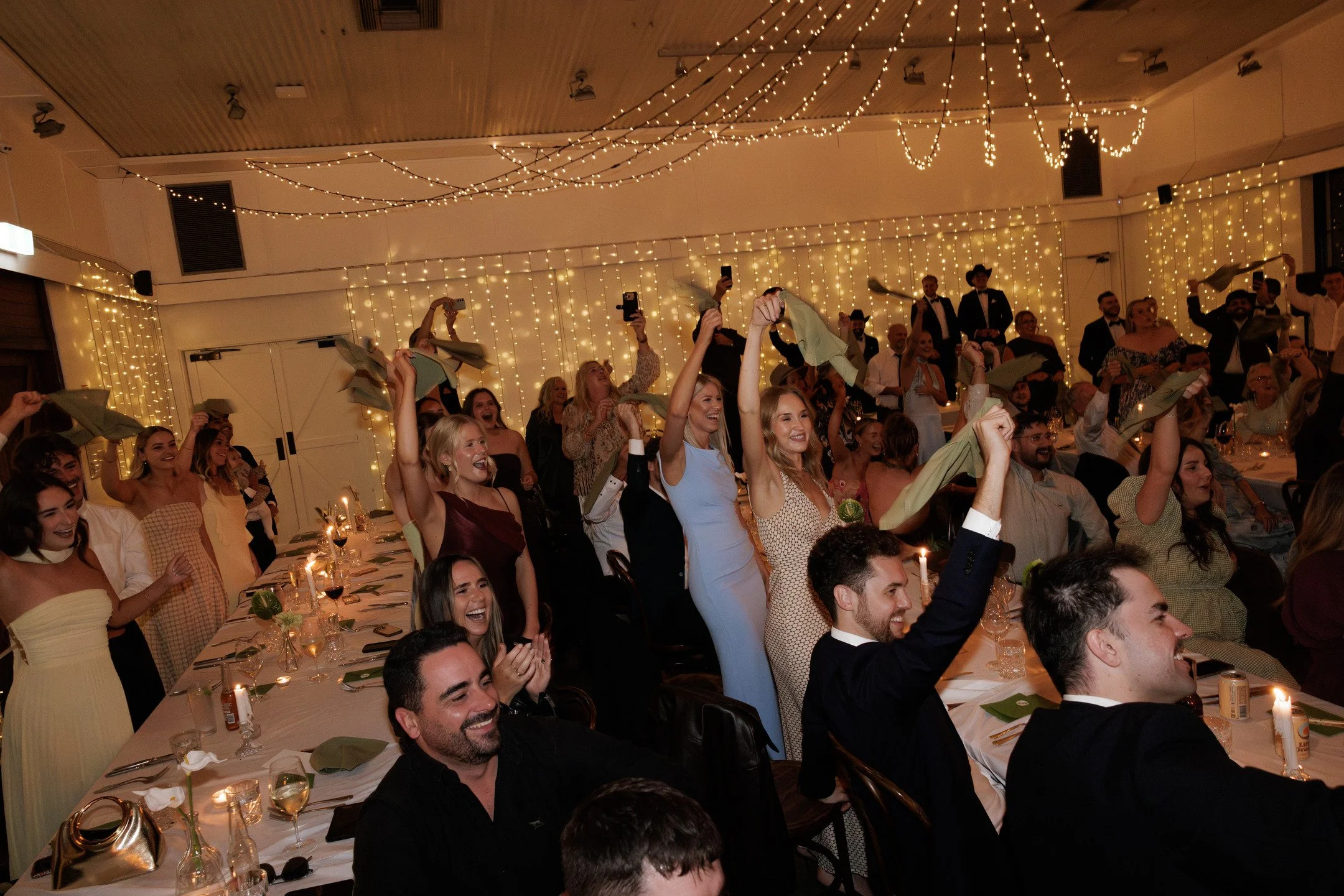 A lively celebration with many people dressed in formal attire, smiling and cheering. The room is decorated with string lights hanging from the ceiling and backdrop, creating a warm, festive atmosphere. The guests are seated at long tables set with d
