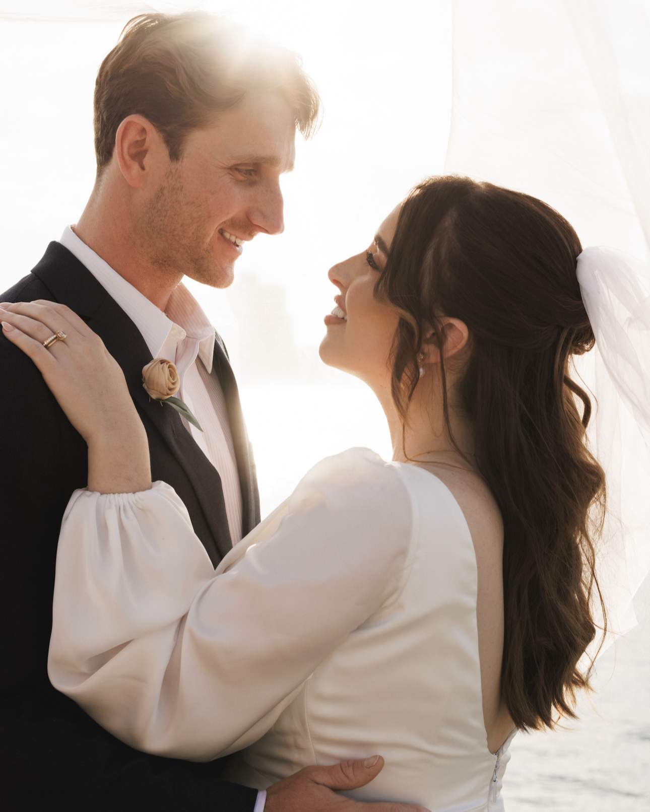 A newlywed couple gazing into each other's eyes indoors, backlit by a bright window, with the bride wearing a white dress and veil, and the groom in a dark suit with a tie.