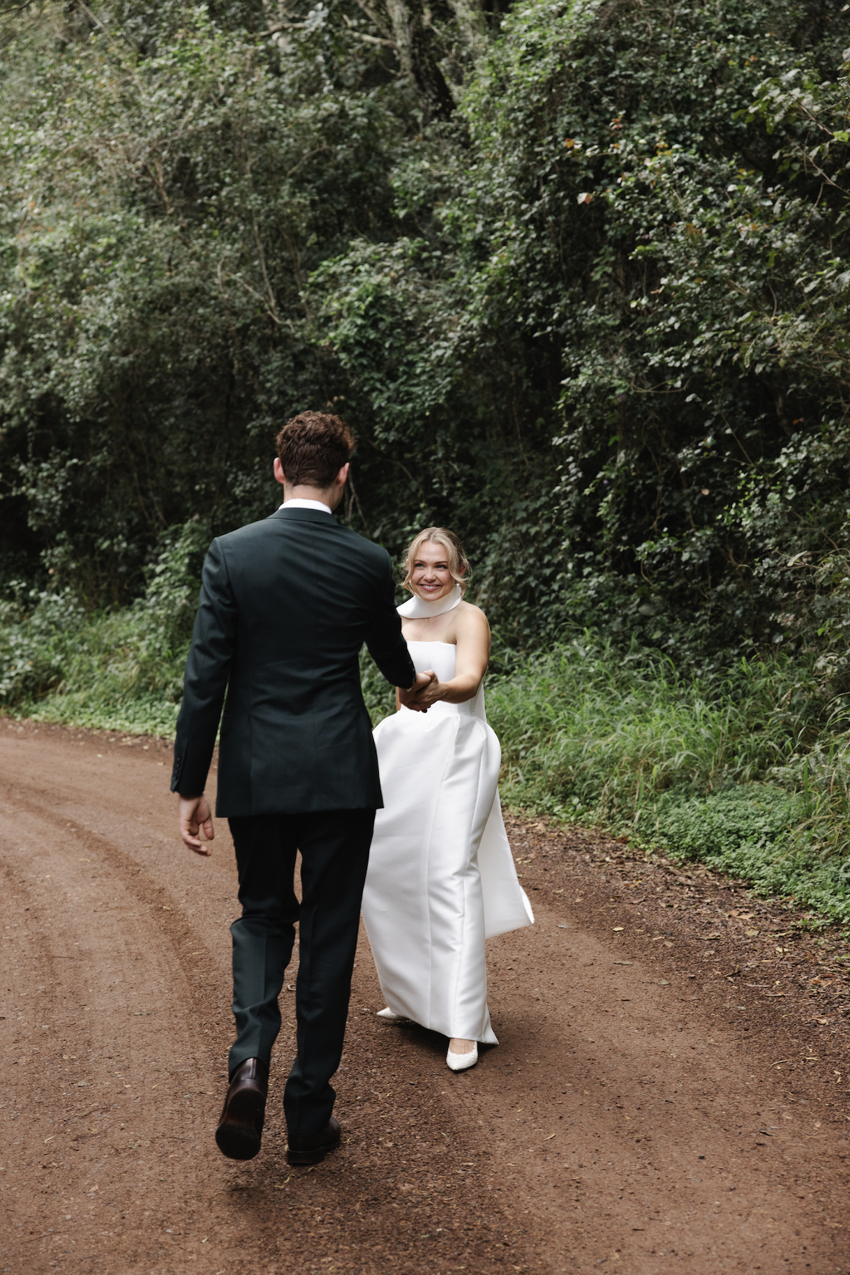A newlywed couple holds hands and walks along a dirt path in a wooded area. The bride smiles at the groom, wearing an elegant white gown, while the groom is dressed in a black suit.