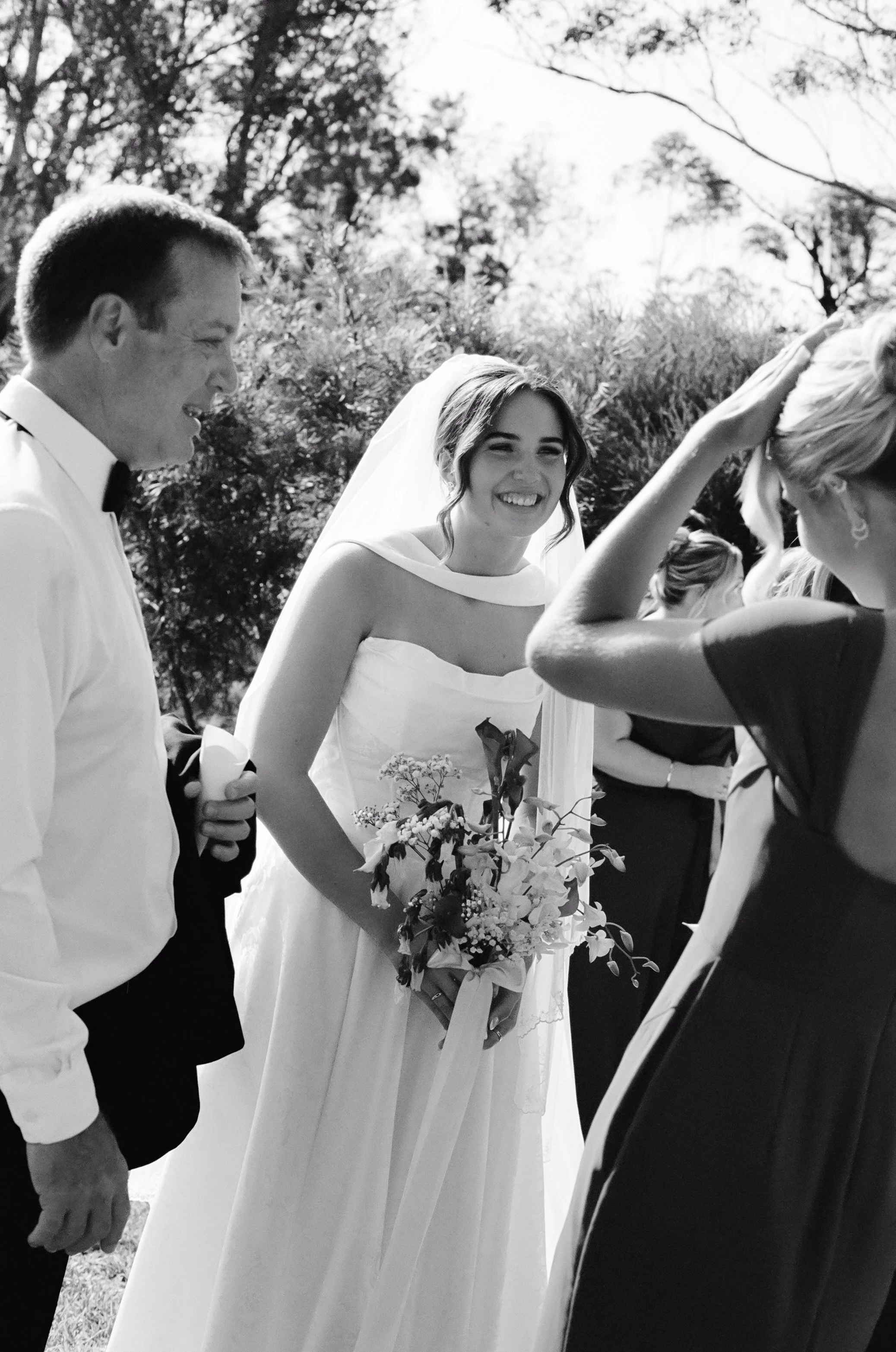Black and white photo of a bride smiling and holding a bouquet, surrounded by wedding guests outdoors.