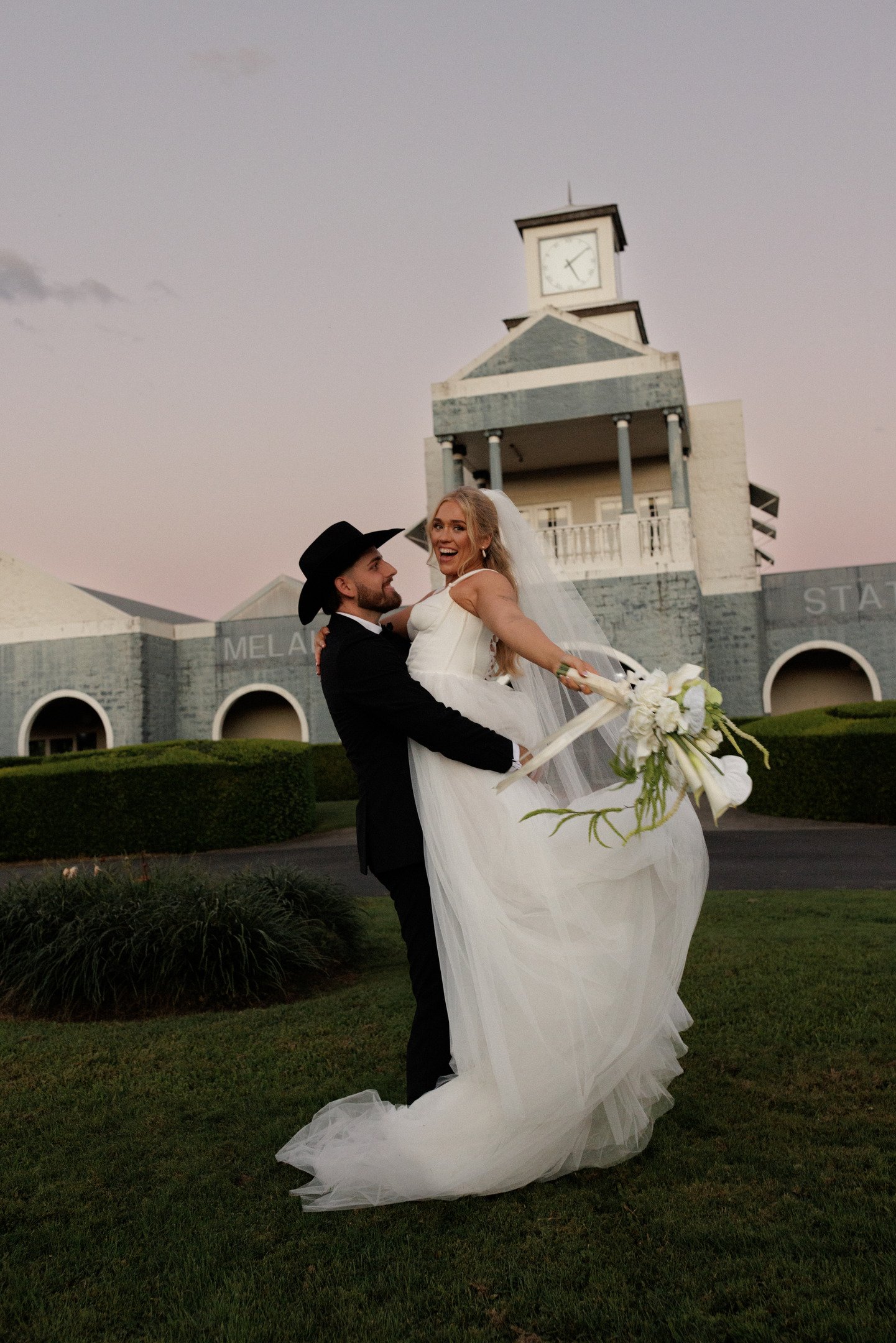 A groom lifting a bride in a wedding dress outside of a building with a clock tower at dusk.