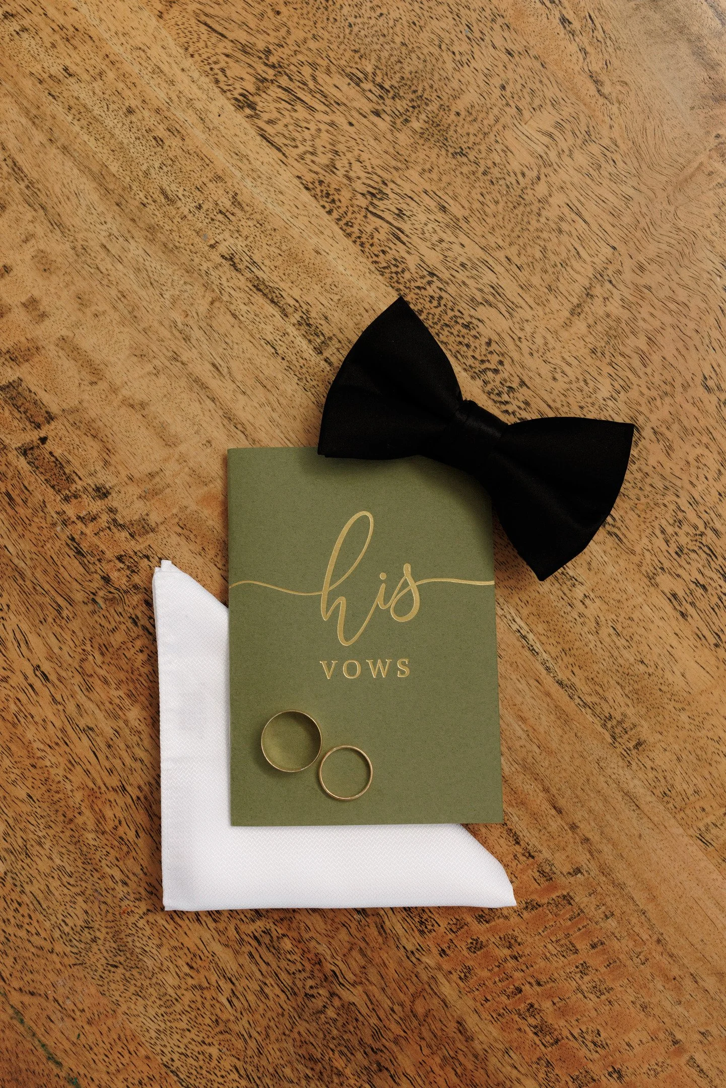 A pair of gold wedding rings on white cloth, with a green card that says 'this vows' and a black bow tie on a wooden surface.