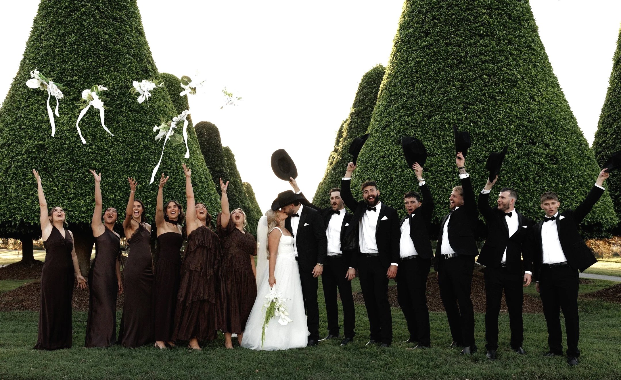 A group of people celebrating outdoors on a sunny day, including women in formal dresses, a bride and groom, and men in tuxedos, with large manicured trees in the background.