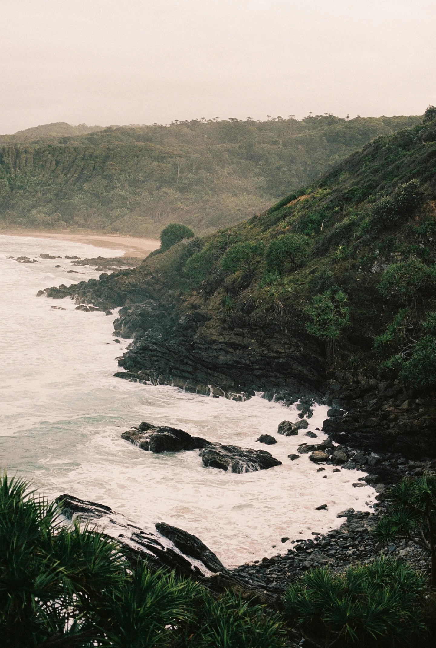 Coastal scene with rocky shoreline, waves hitting rocks, green hillside covered in trees and bushes, overcast sky.