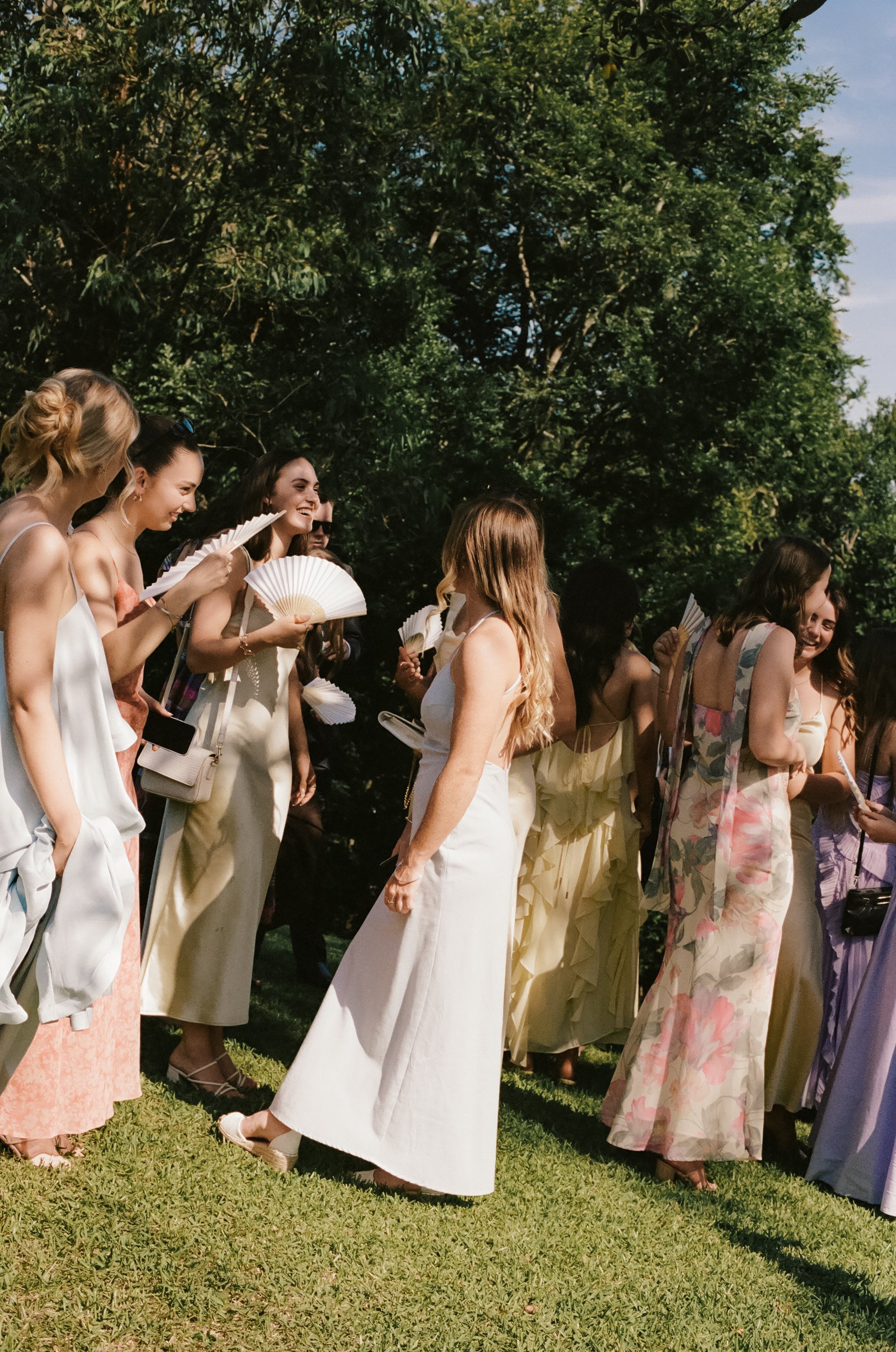 A group of women dressed in summer dresses, holding fans, gathered outdoors on a sunny day with green trees in the background.