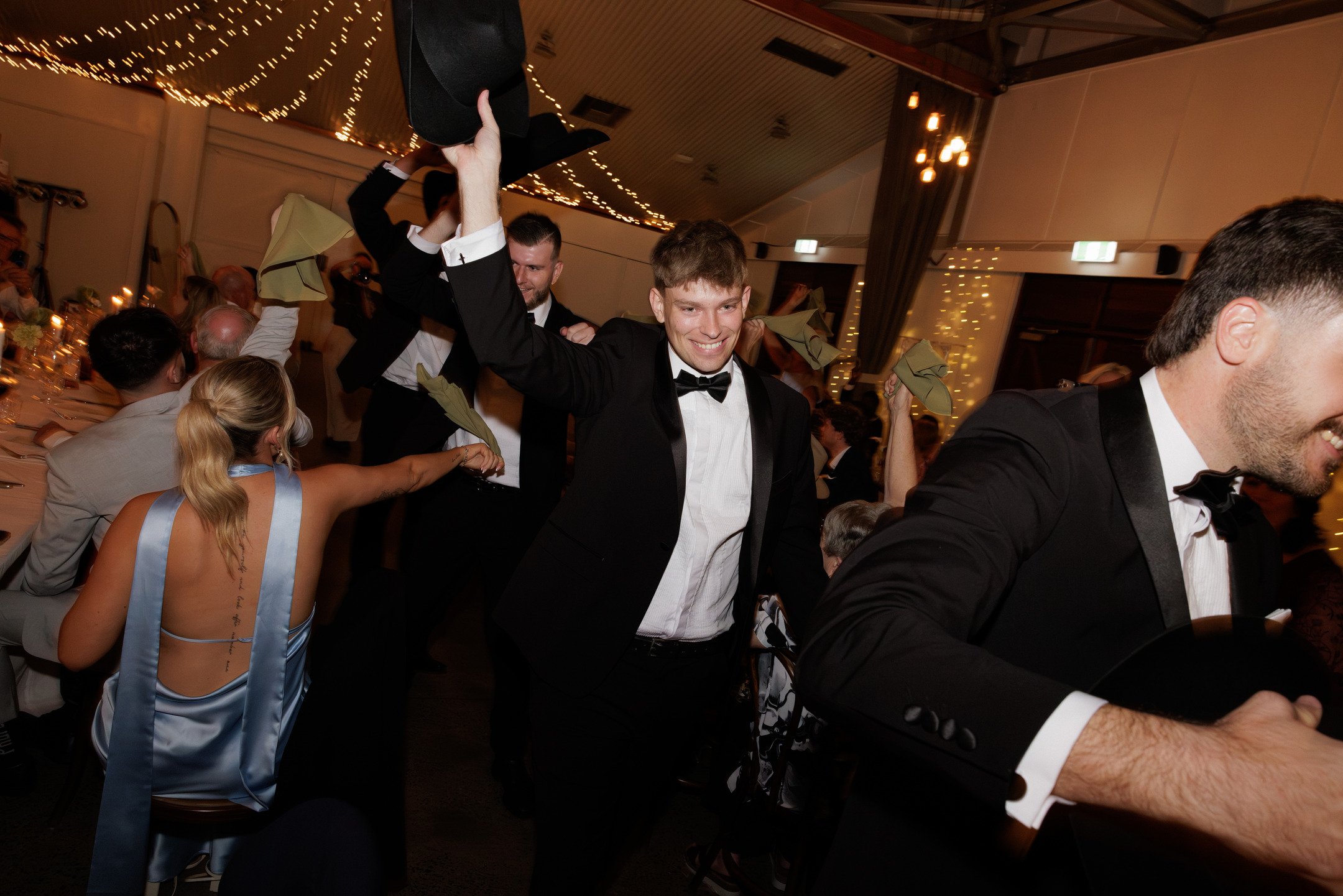 Young men in tuxedos dancing at a formal event, with some raising their hats in celebration, surrounded by seated guests in elegant attire inside a decorated banquet hall.
