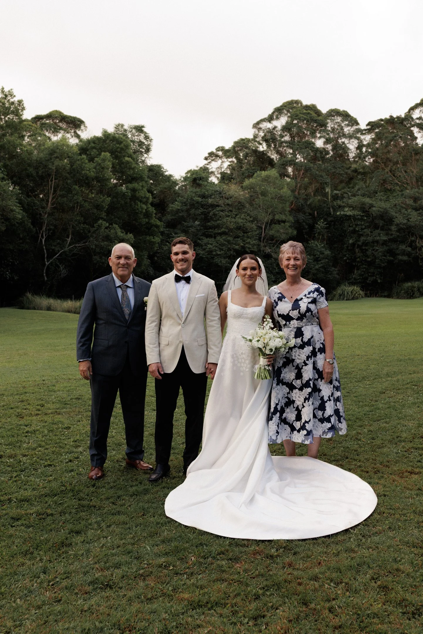 A wedding party with four people standing on a grassy field with a background of tall trees. The bride is dressed in a white gown holding a bouquet, flanked by two men in formal suits, and a woman in a floral dress.