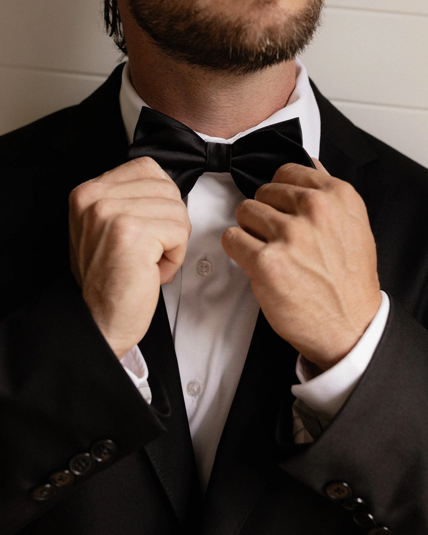 A person wearing a black tuxedo with a white dress shirt, adjusting a black bow tie with both hands.