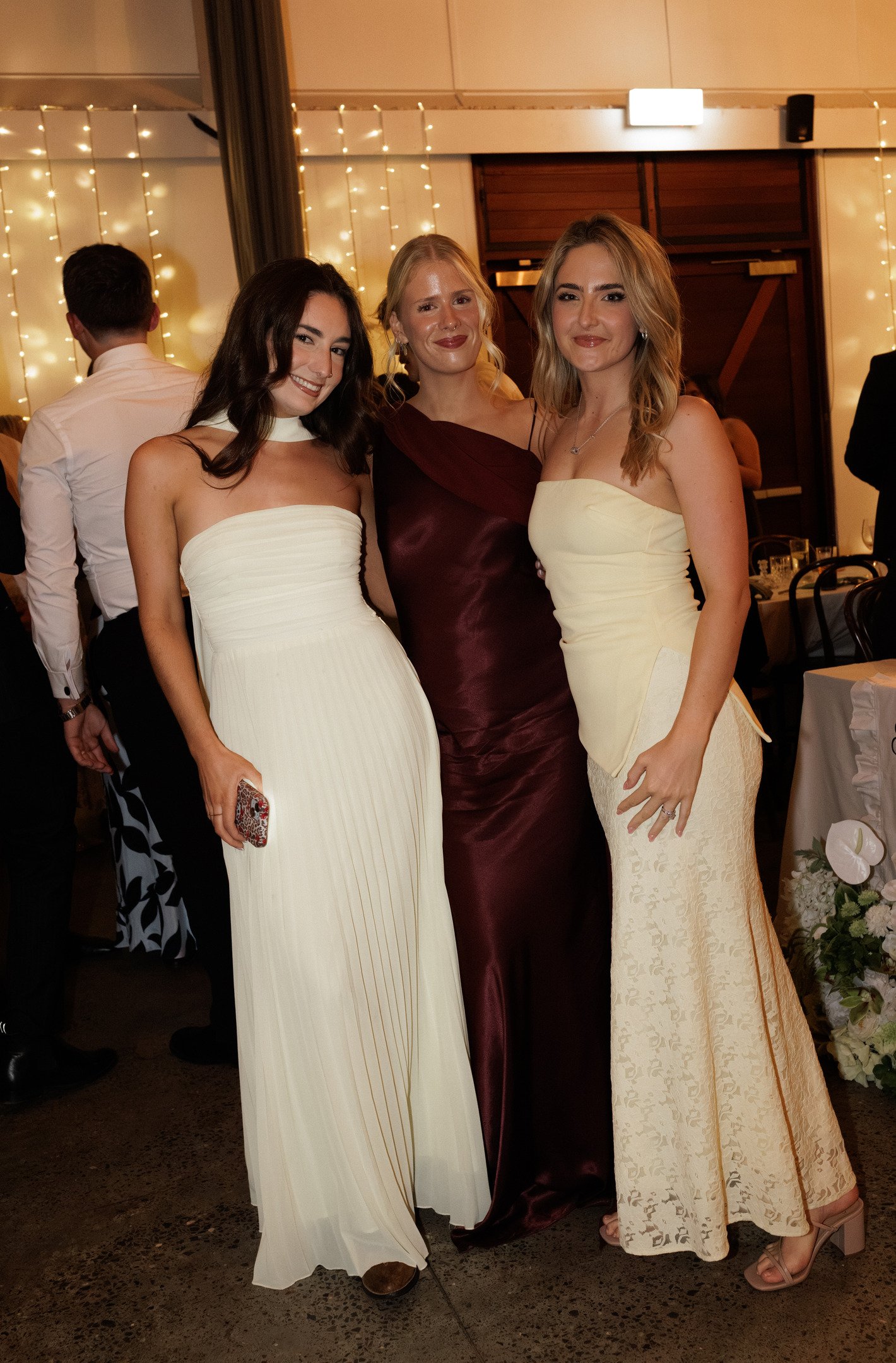 Three women dressed in formal gowns at an indoor event, smiling for the camera, with decorated curtains and tables in the background.