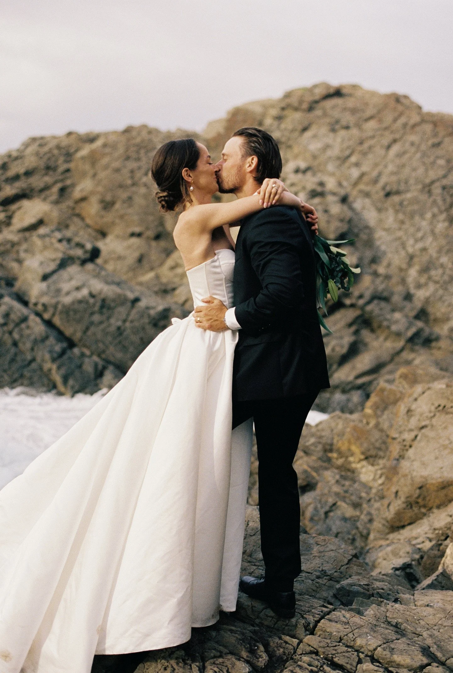 A bride and groom sharing a kiss on a rocky beach at sunset, with rocky cliffs in the background.