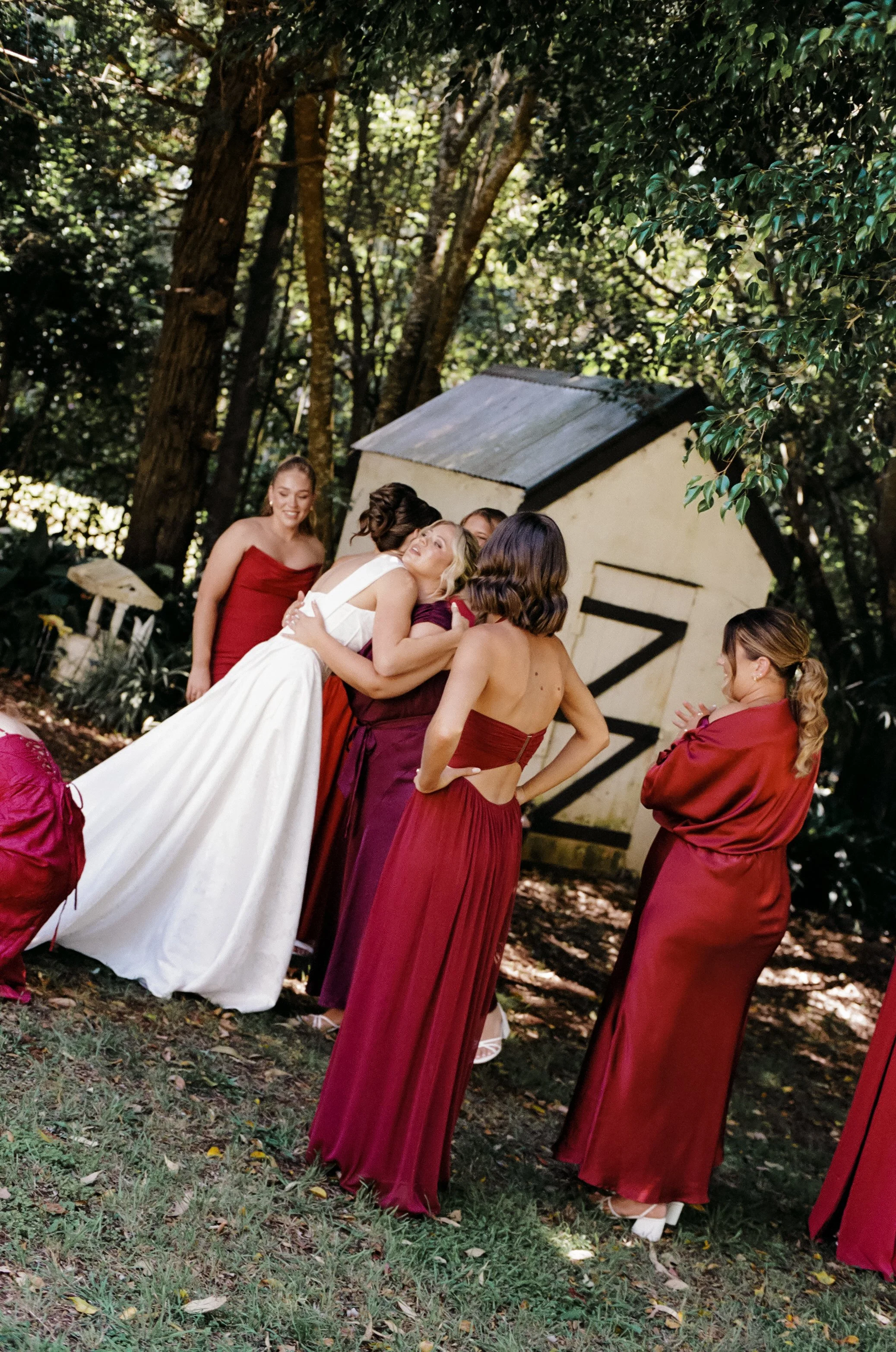 Group of women in burgundy dresses hugging and smiling outdoors near a small shed surrounded by trees.