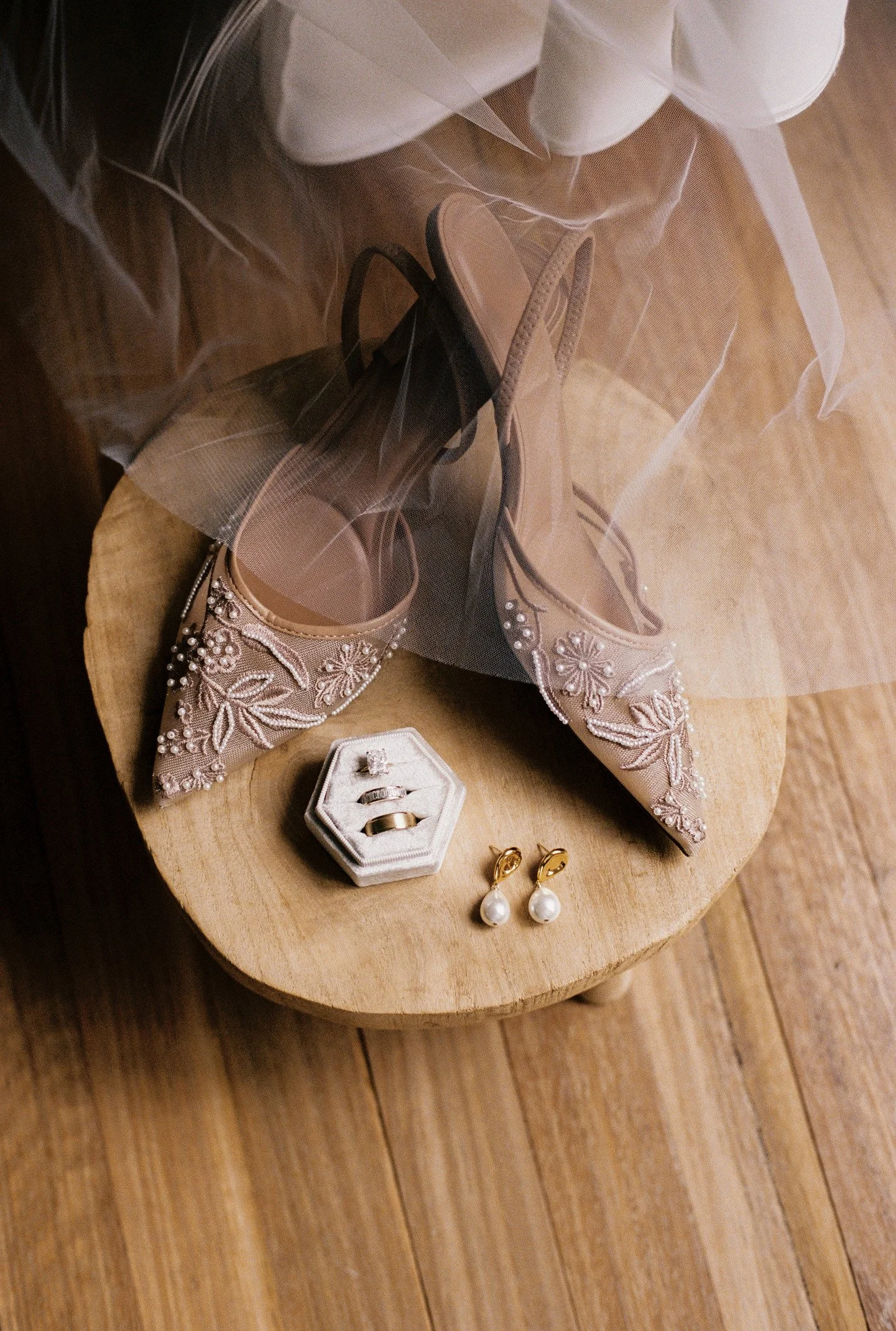 Array of jewelry including pearl earrings, a ring, and a bracelet, along with a pair of nude-colored heels with embellishments and a black high heel, all on a wooden surface.