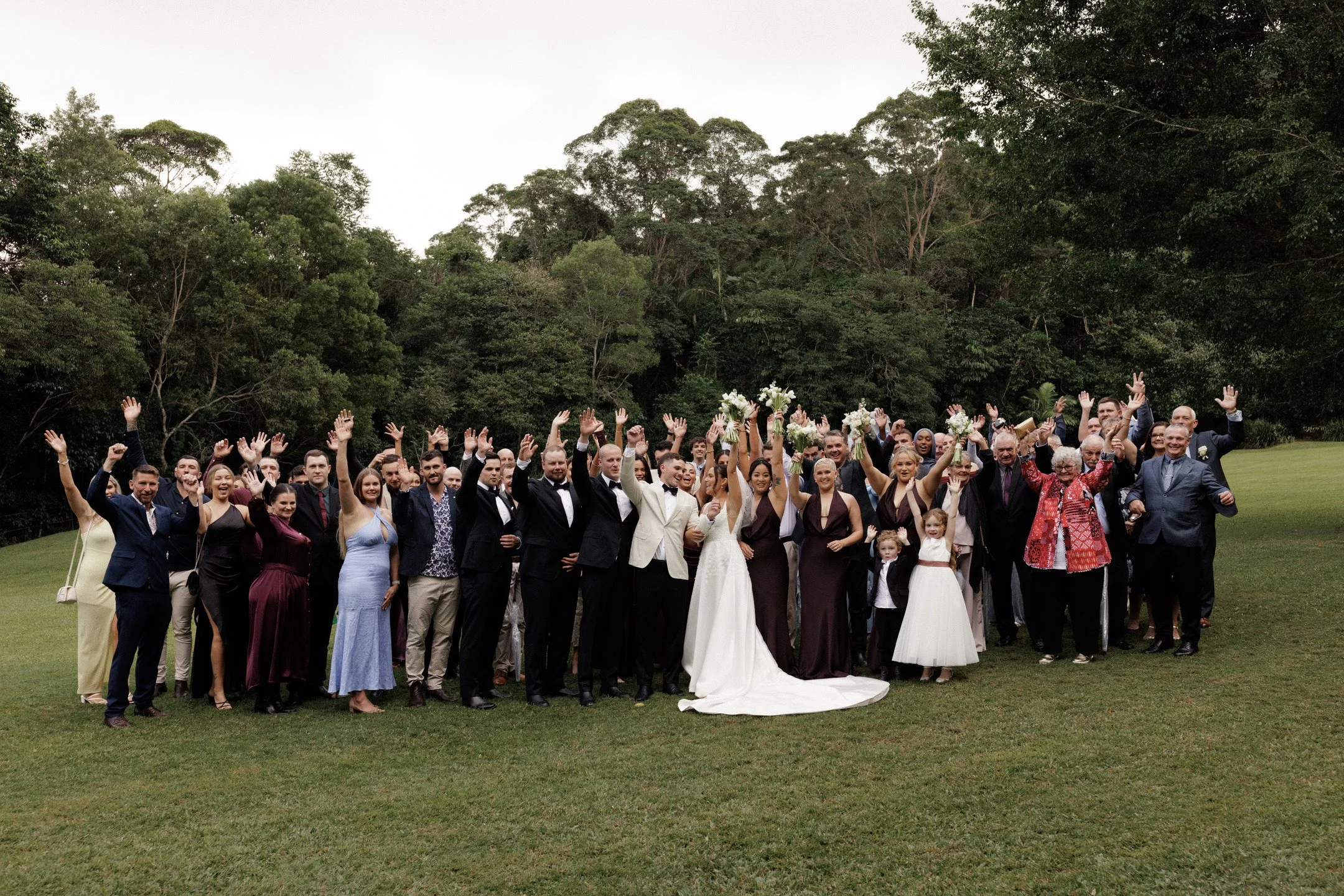 A large group of people dressed in formal attire celebrating outdoors, with some holding bouquets of white flowers, in front of a lush green backdrop of trees.