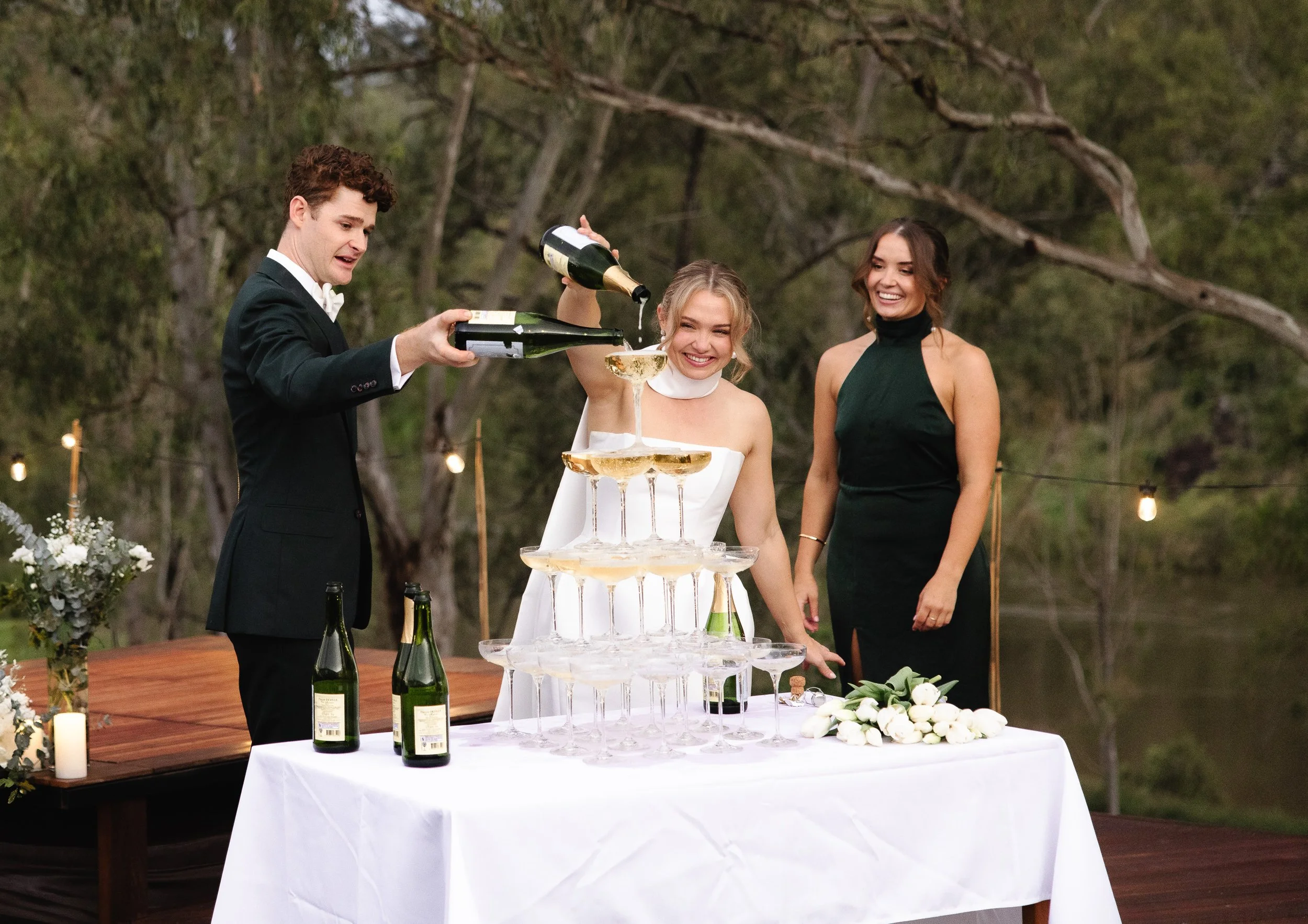 A man in a tuxedo pours champagne into a tower of glasses, with a smiling woman in a white dress and another woman in a black dress watching during an outdoor celebration at sunset.