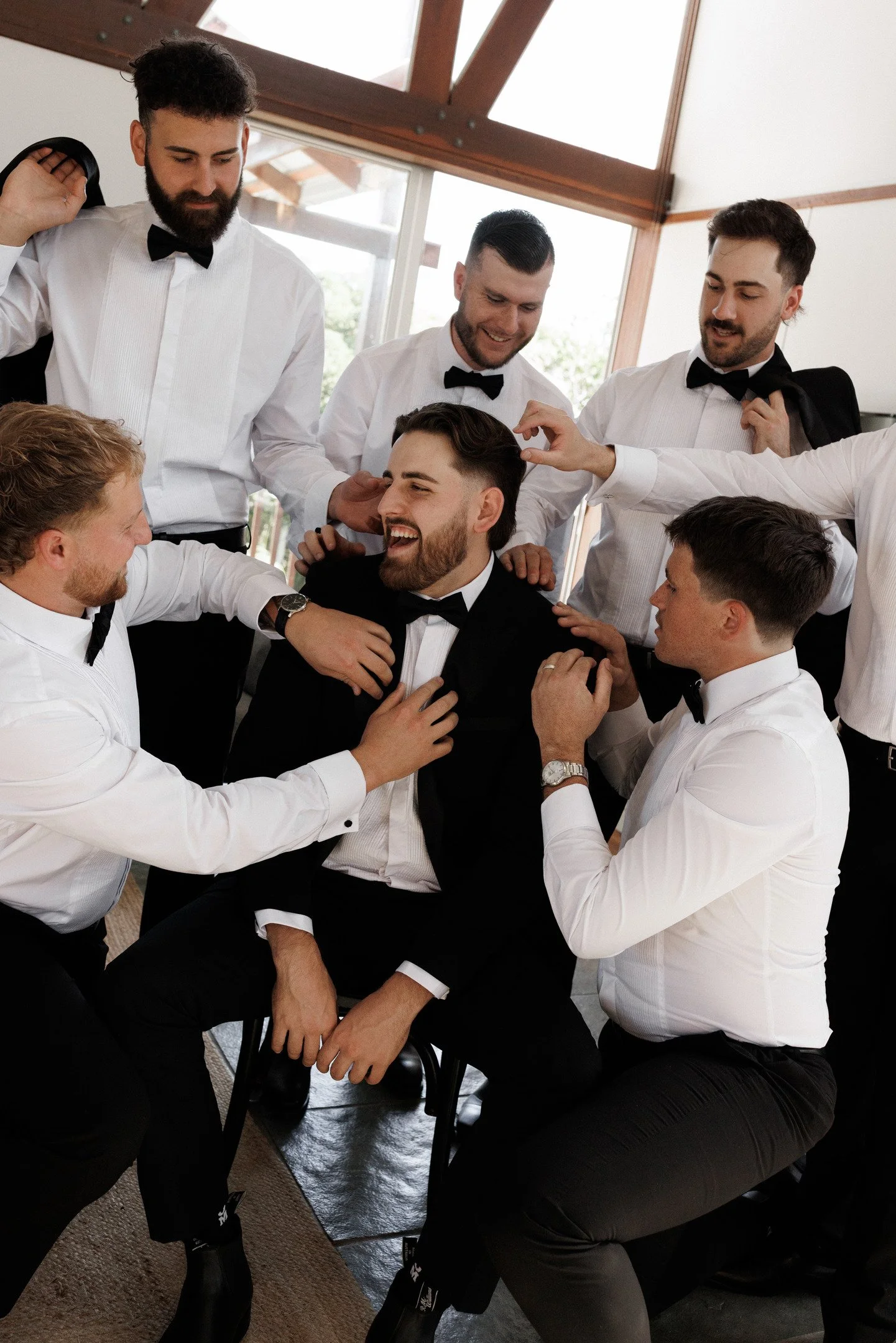 A group of men dressed in white shirts, black tuxedos, and black bow ties, gathered around a seated man in a tuxedo, smiling and interacting cheerfully inside a room with large windows and wooden beams.