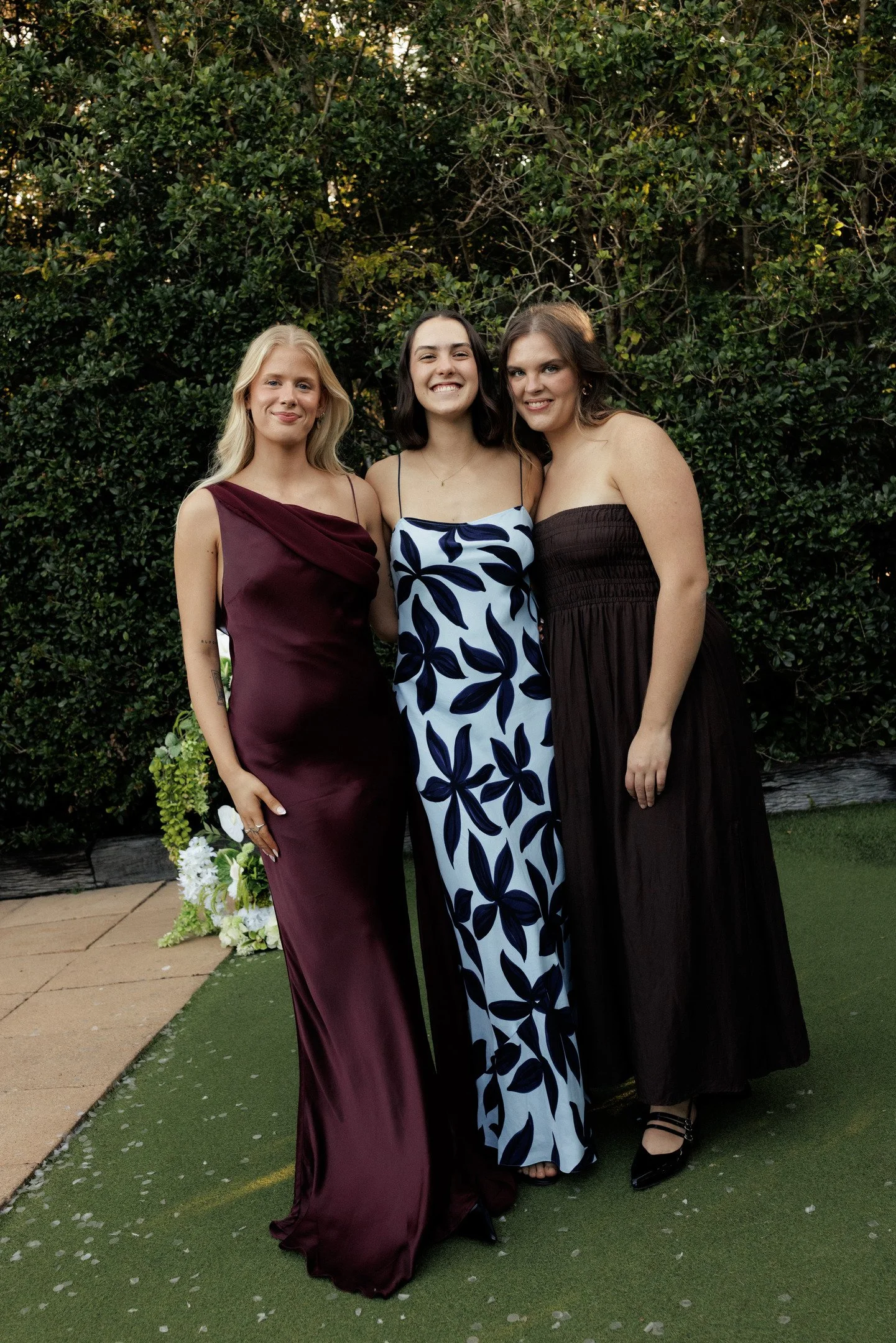Three women standing close together outdoors in front of a dense green hedge, dressed in formal dresses, smiling and posing for the camera.