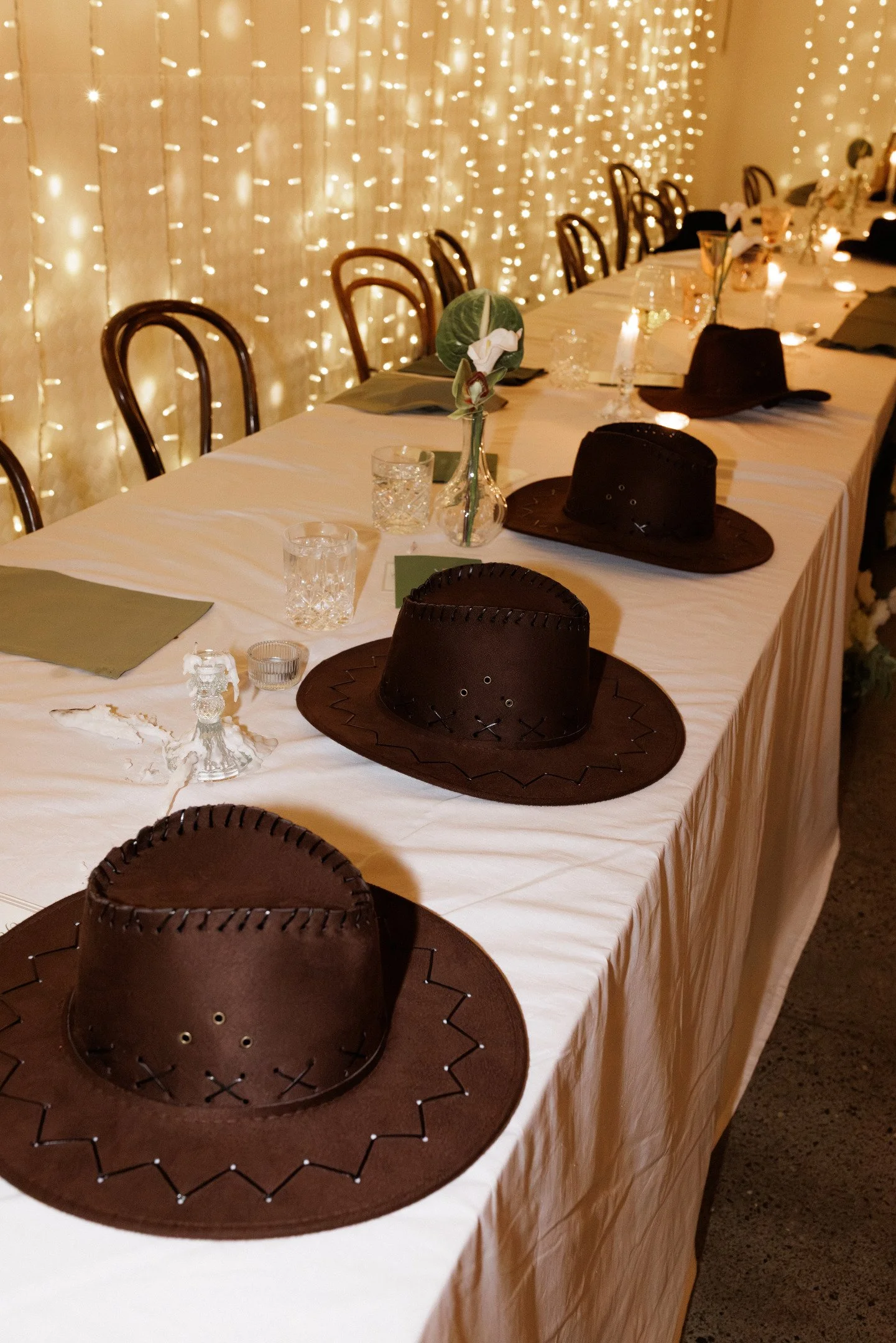 A long banquet table decorated for a Western-themed event, featuring cowboy hats, glassware, and floral centerpieces, illuminated by string lights in the background.
