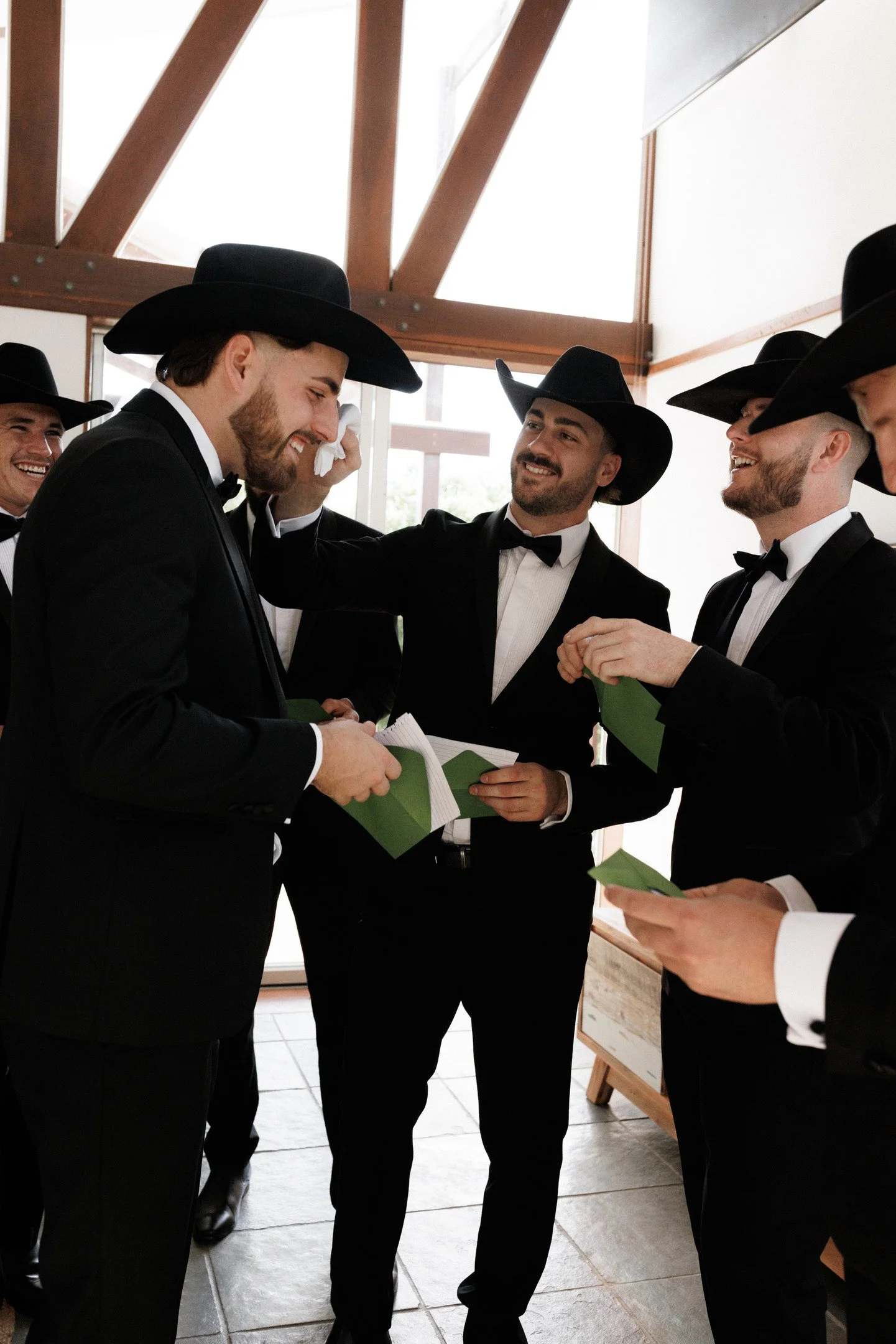 Group of men in tuxedos and cowboy hats smiling and talking indoors, holding green cards or papers.