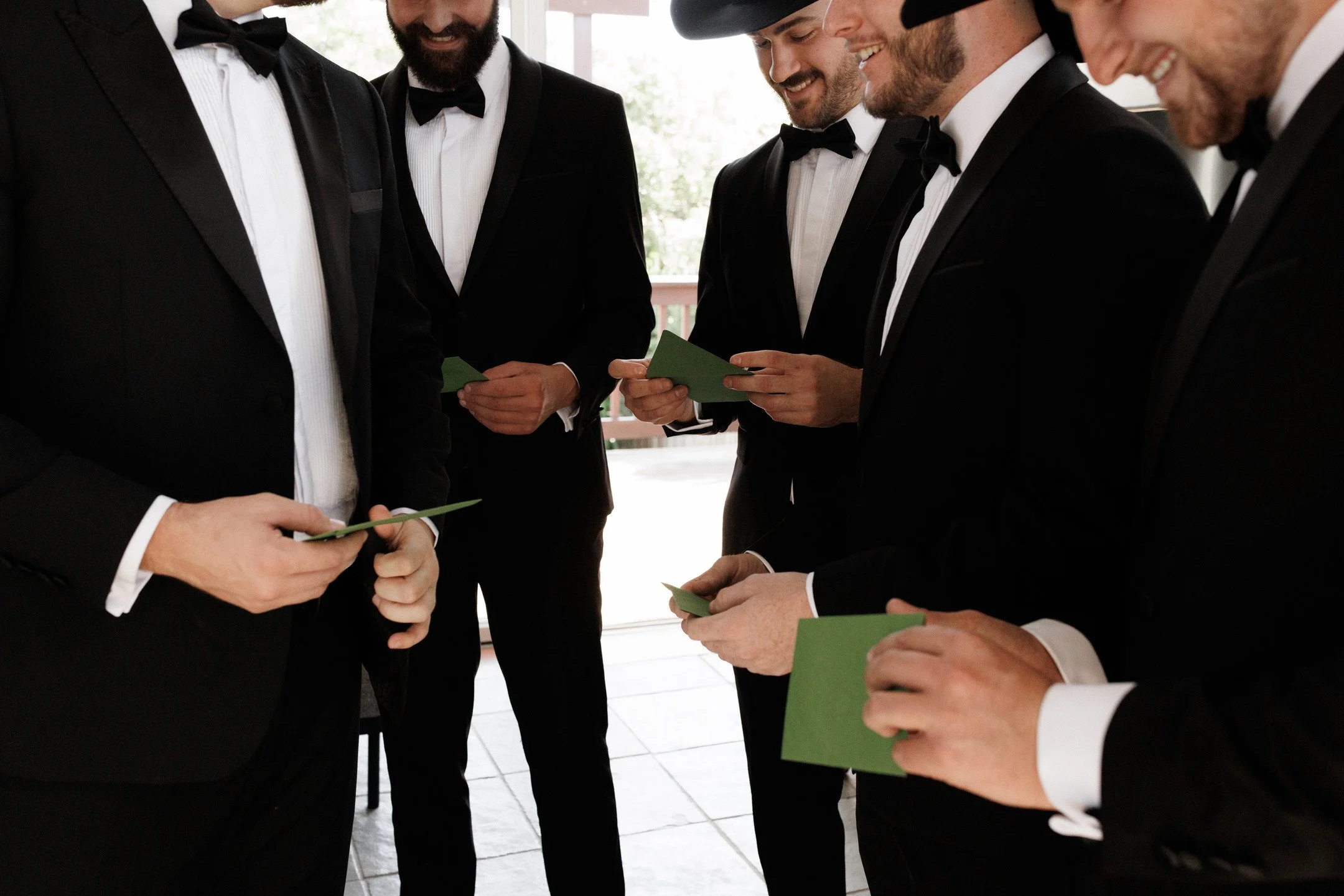 A group of men wearing tuxedos and bow ties holding green cards and smiling at an indoor event.