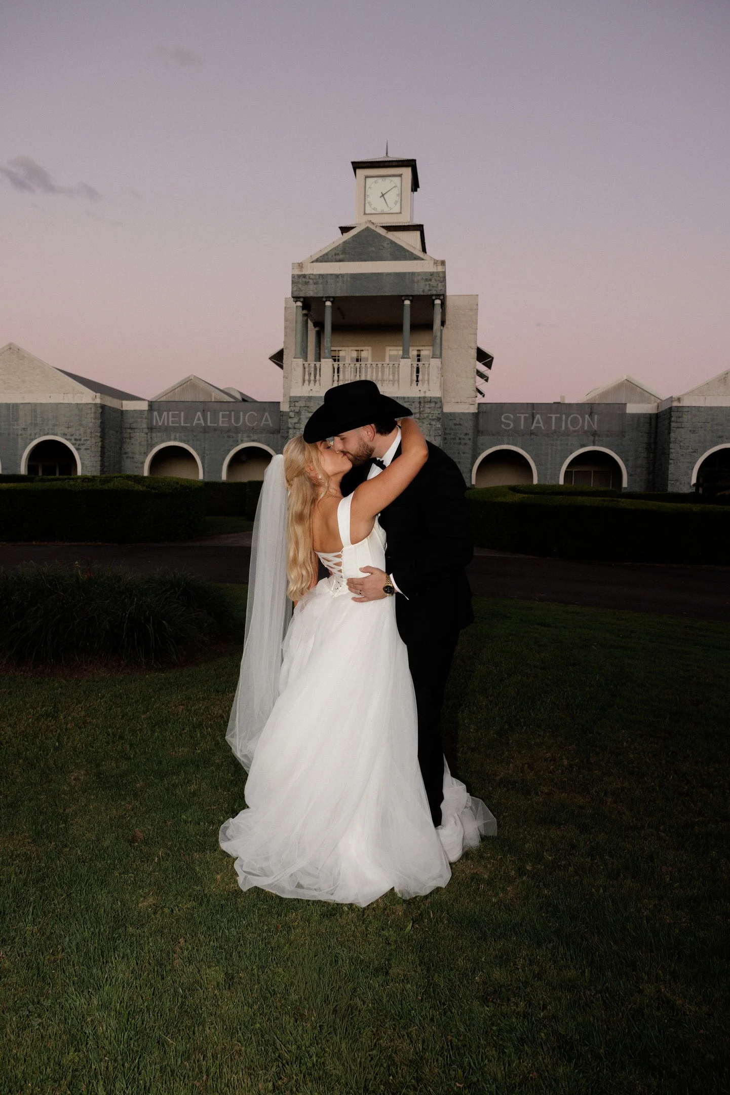 A bride and groom sharing a kiss in front of a historic train station at dusk. The bride is in a white wedding gown and the groom in a black tuxedo. The station has a clock showing about 7:09.