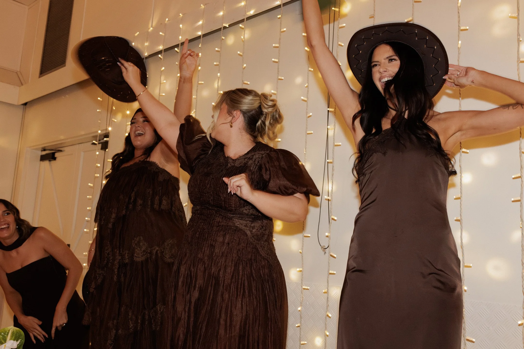 Group of women celebrating at an indoor party, wearing dark dresses and black hats, with string lights in the background.