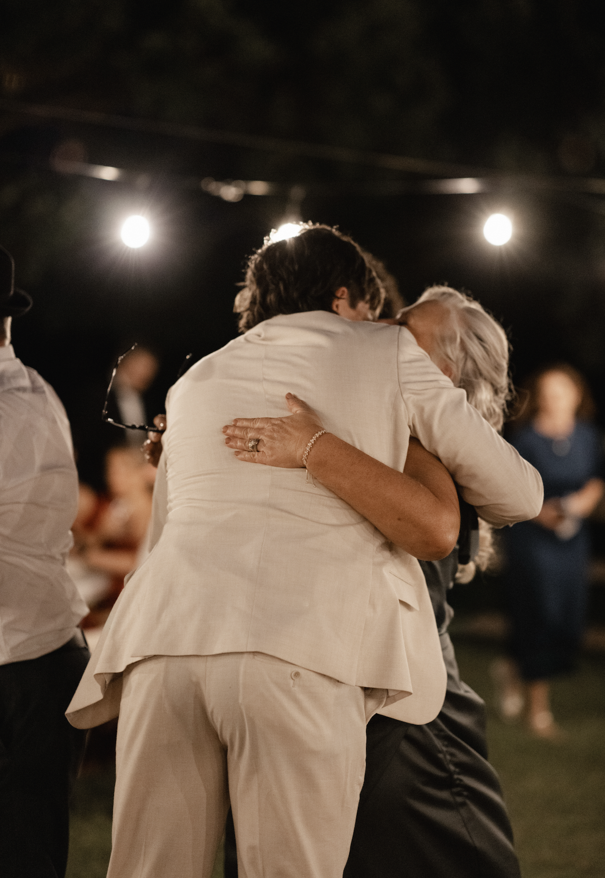 An elderly woman and a middle-aged man embrace in a dance or hug at a social gathering indoors, with other people and bright lights in the background.