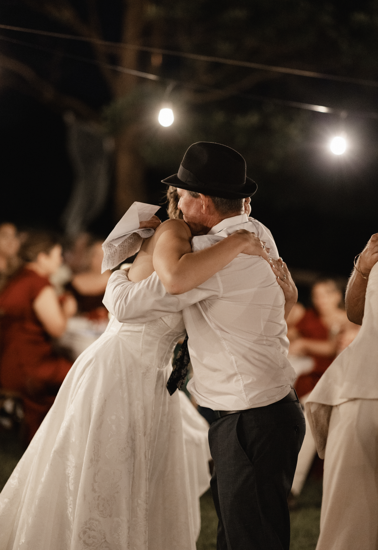 Two people hugging at an outdoor event, one wearing a white dress and the other a white shirt with a black hat, under string lights at night.