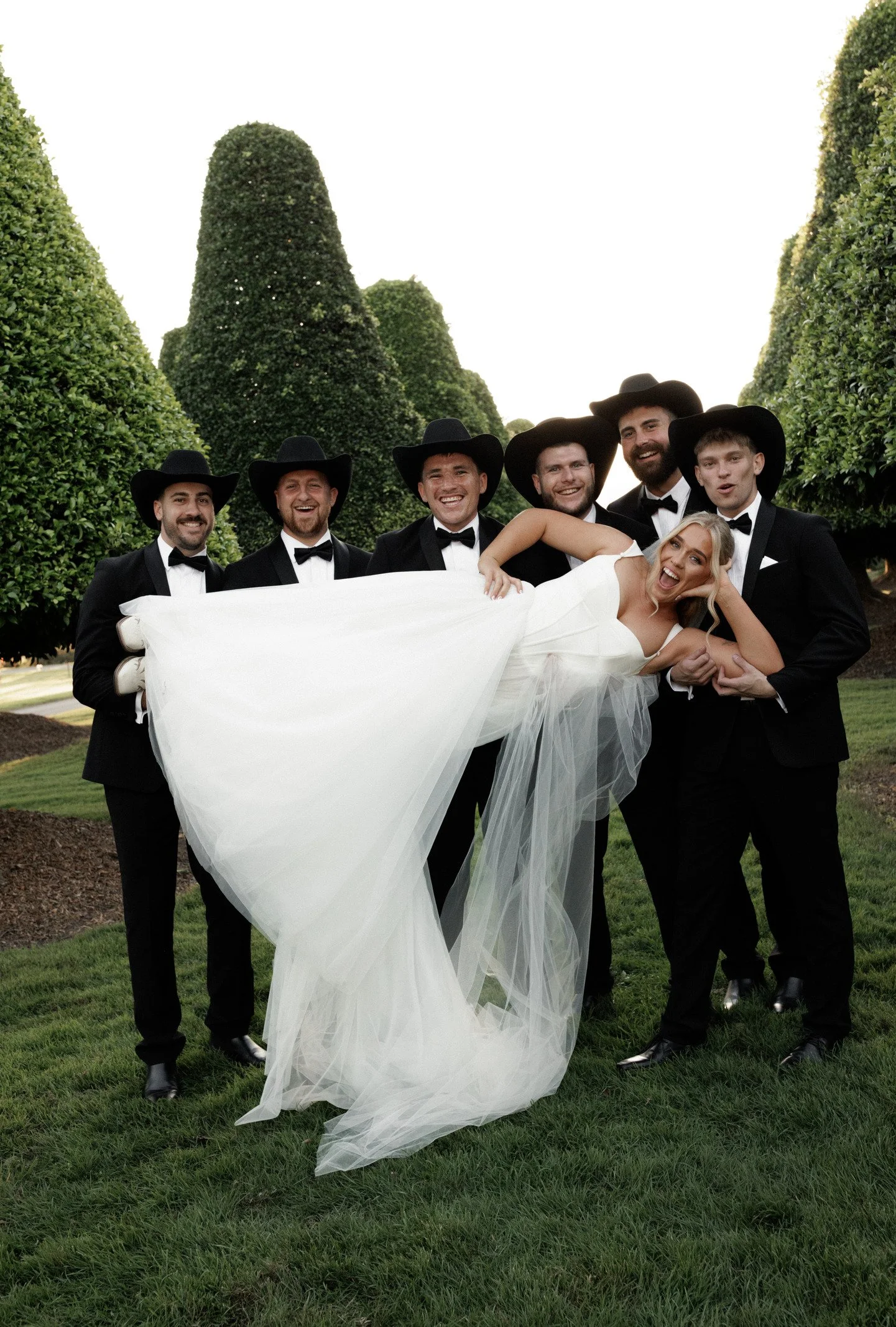 Group of seven men and a woman dressed in formal wedding attire outside on green grass, with large, trimmed bushes in the background. The women, wearing a white wedding dress, is being held horizontally by the men, who are dressed in black tuxedos wi