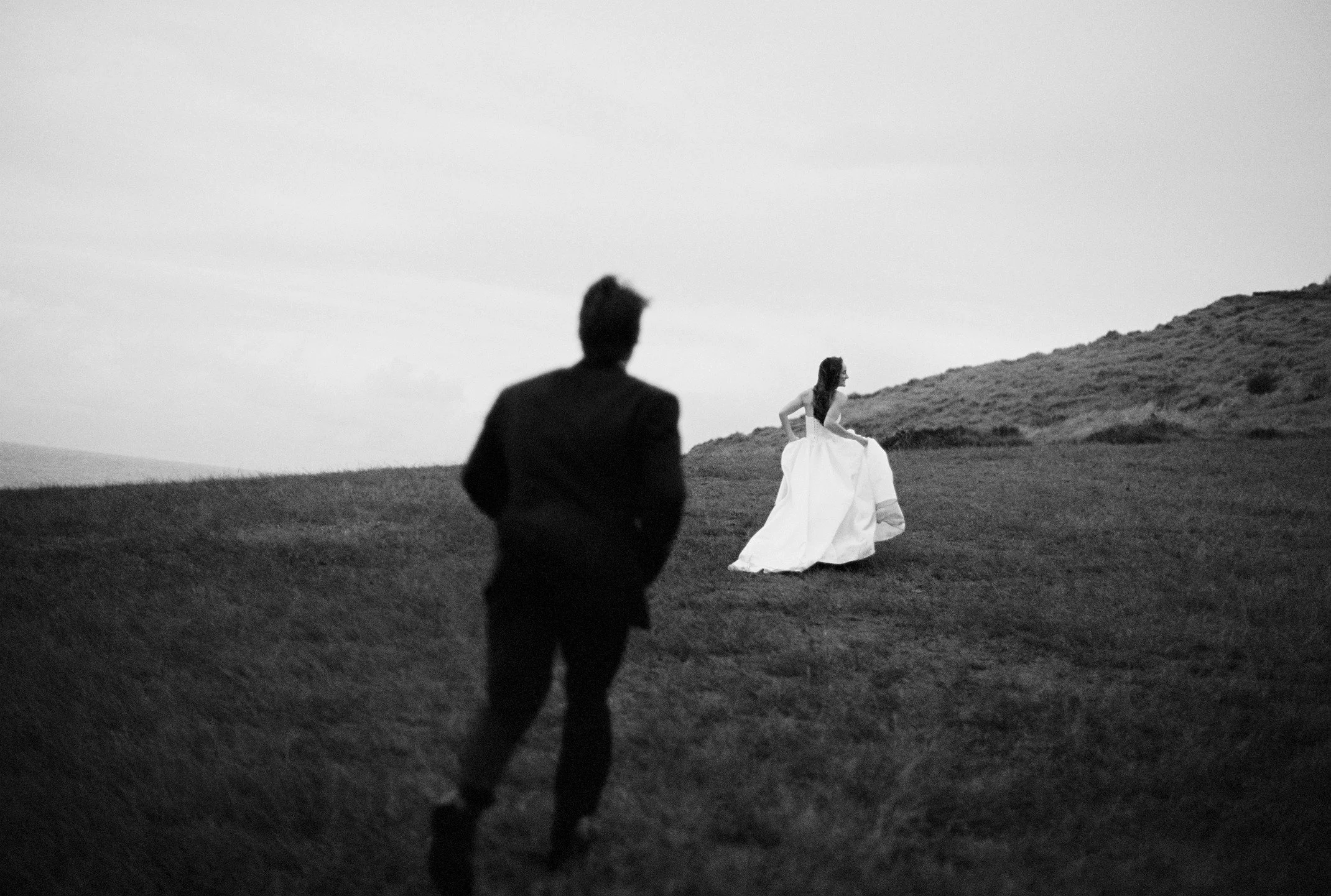 A black and white photo of a woman in a long dress walking along a grassy hillside with two men following behind her.