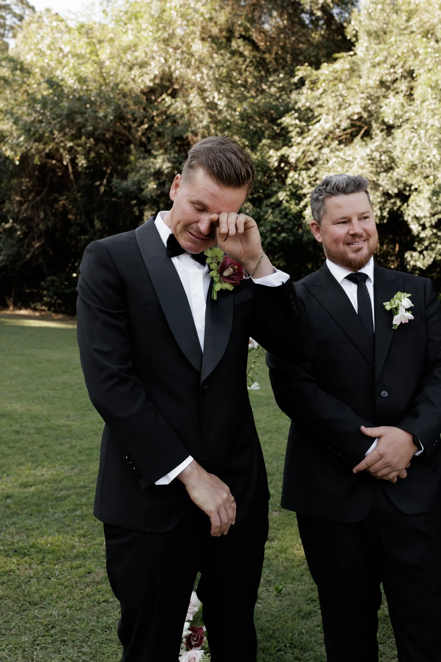 Two men in black tuxedos with white shirts and bow ties standing outdoors on grass with trees in the background. One man is emotional, wiping his eye, while the other smiles gently. Each has a white boutonniere and a floral arrangement on their tuxed