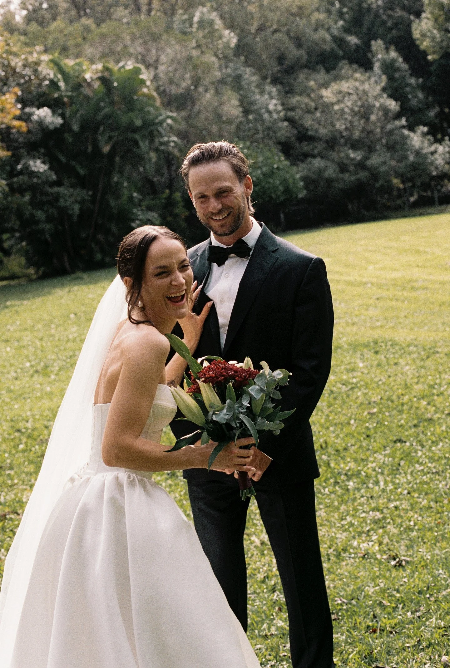 A smiling bride and groom standing outdoors on a grassy area, with the bride holding a bouquet of flowers and the groom in a tuxedo. The background features trees and sunlight.