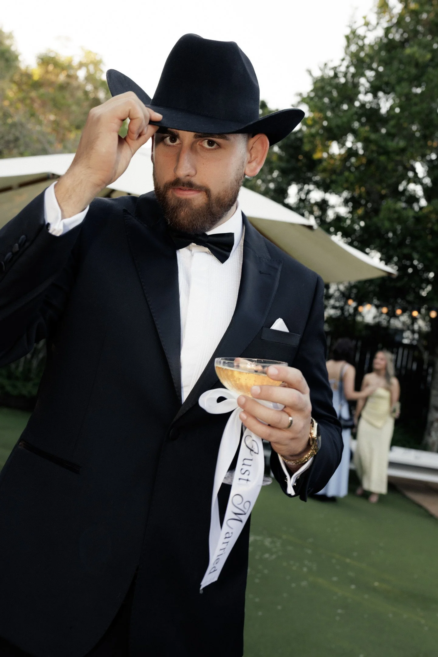 A man dressed in a tuxedo and black cowboy hat holding a glass of champagne and a white sash with text, standing outside at a formal event with people and trees in the background.